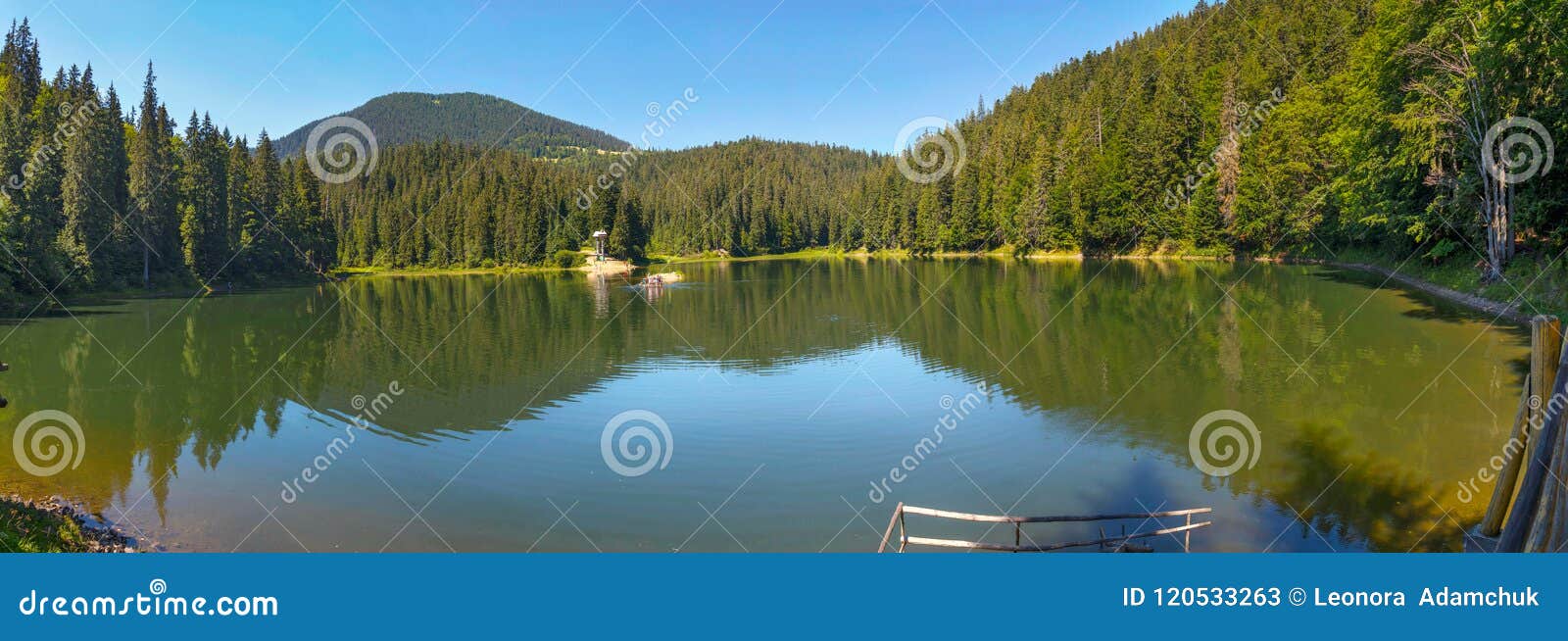 Green Lake In The Mountains Among The Coniferous Forest Under The Blue