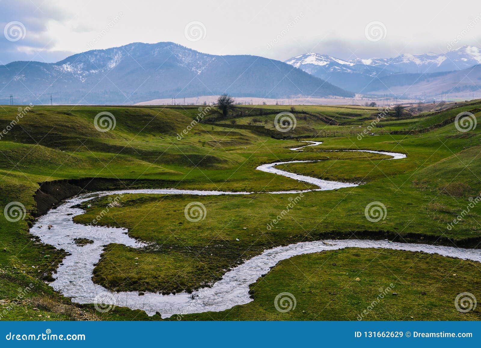 Green Meadow With A Small River Against The Background Of Snow Capped