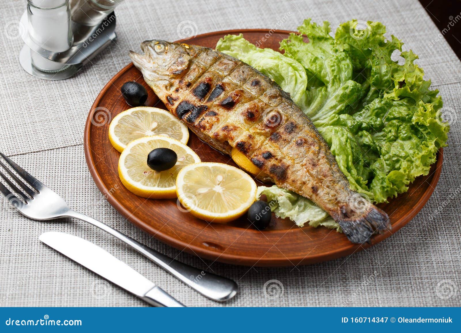 grilled trout in brown clay bowl close-up. front view of plate with grilled trout garnishe with salade, lemon and olives. focus on