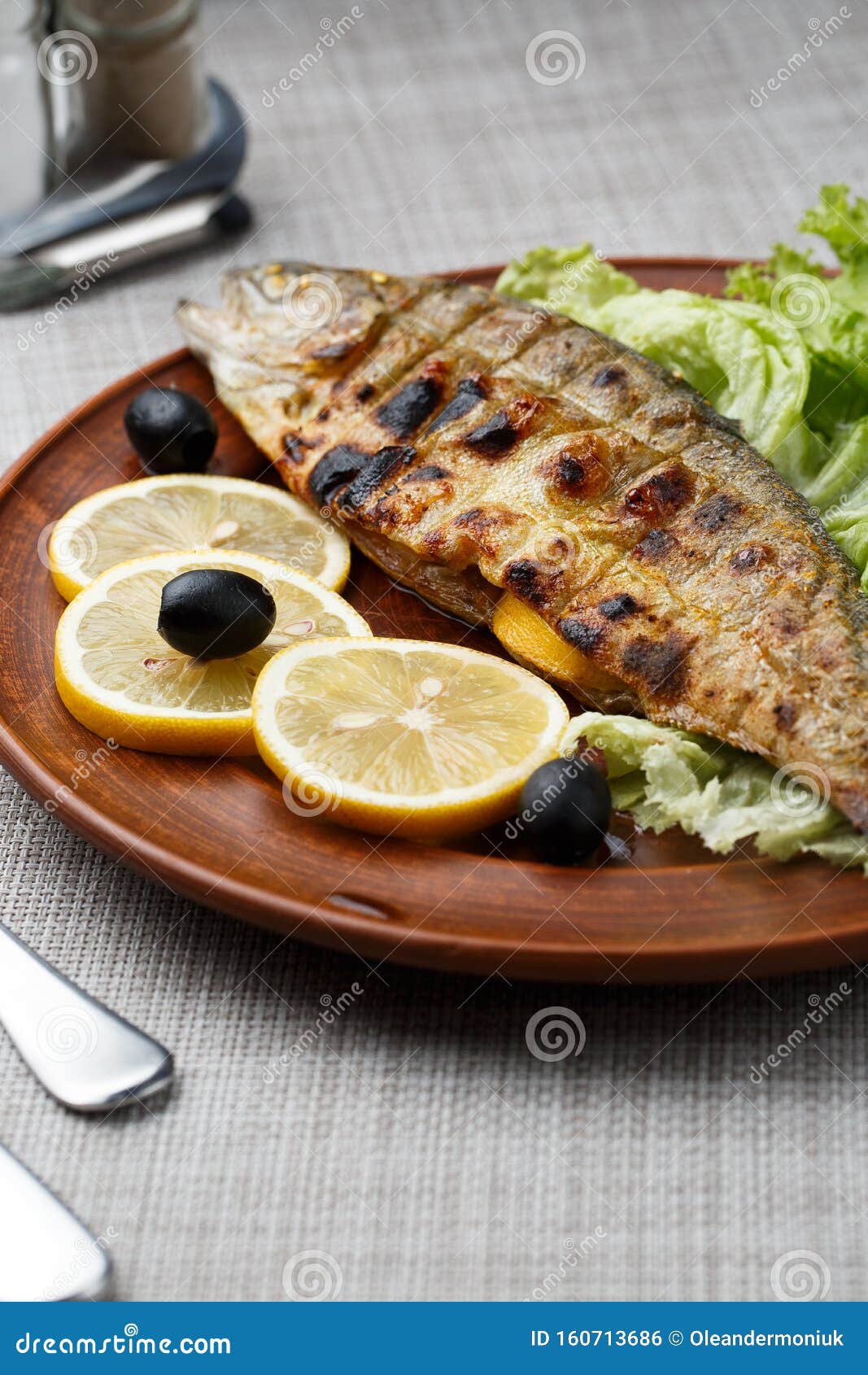 grilled trout in brown clay bowl close-up. front view of plate with grilled trout garnishe with salade, lemon and olives. focus on