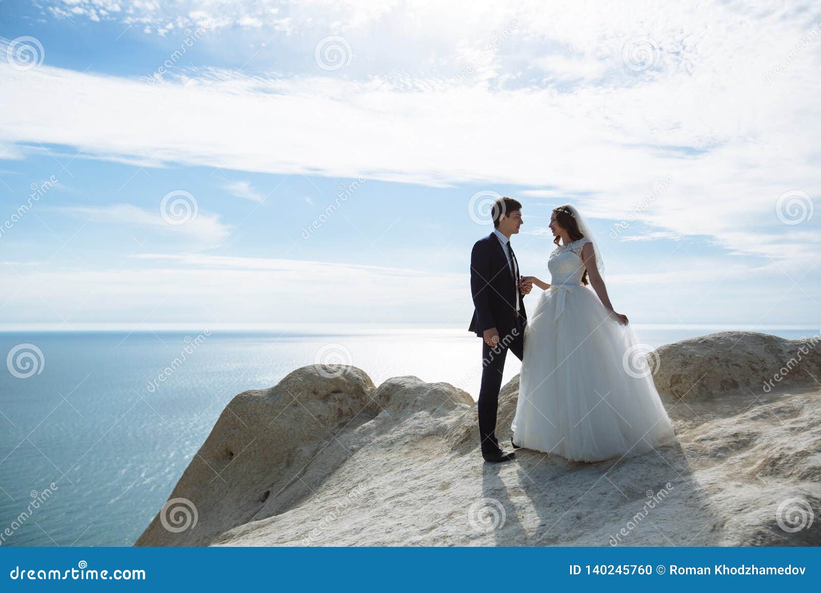 groom in elegant suit and bride in white dress at weddingday on cliff with beautiful view of ocean