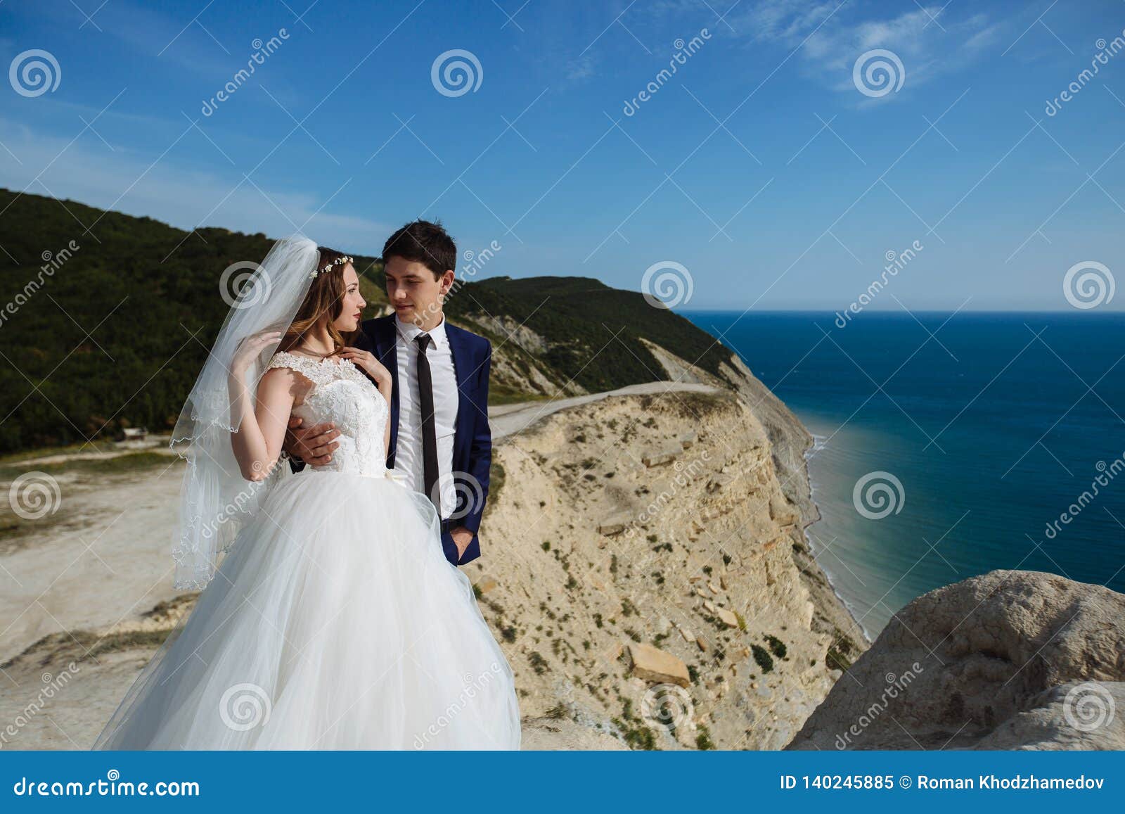 groom in elegant suit and bride in white dress at weddingday on cliff with beautiful view of ocean