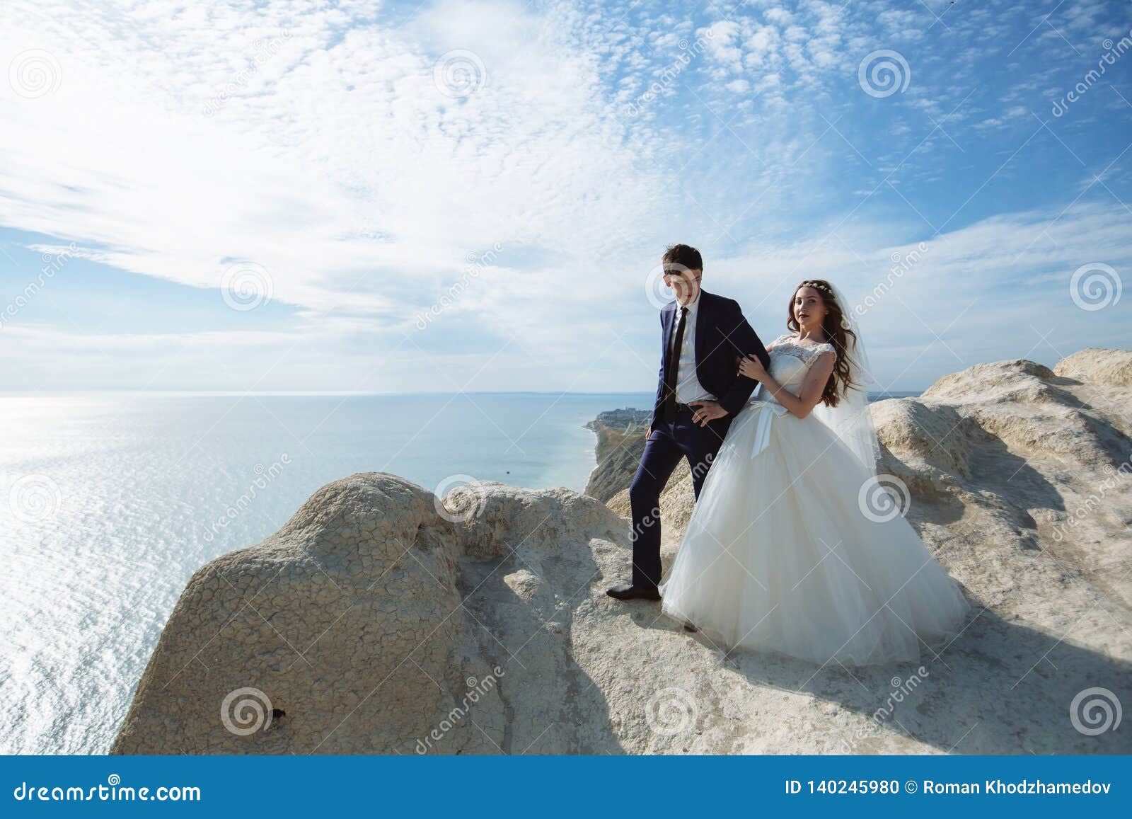 groom in elegant suit and bride in white dress at weddingday on cliff with beautiful view of ocean