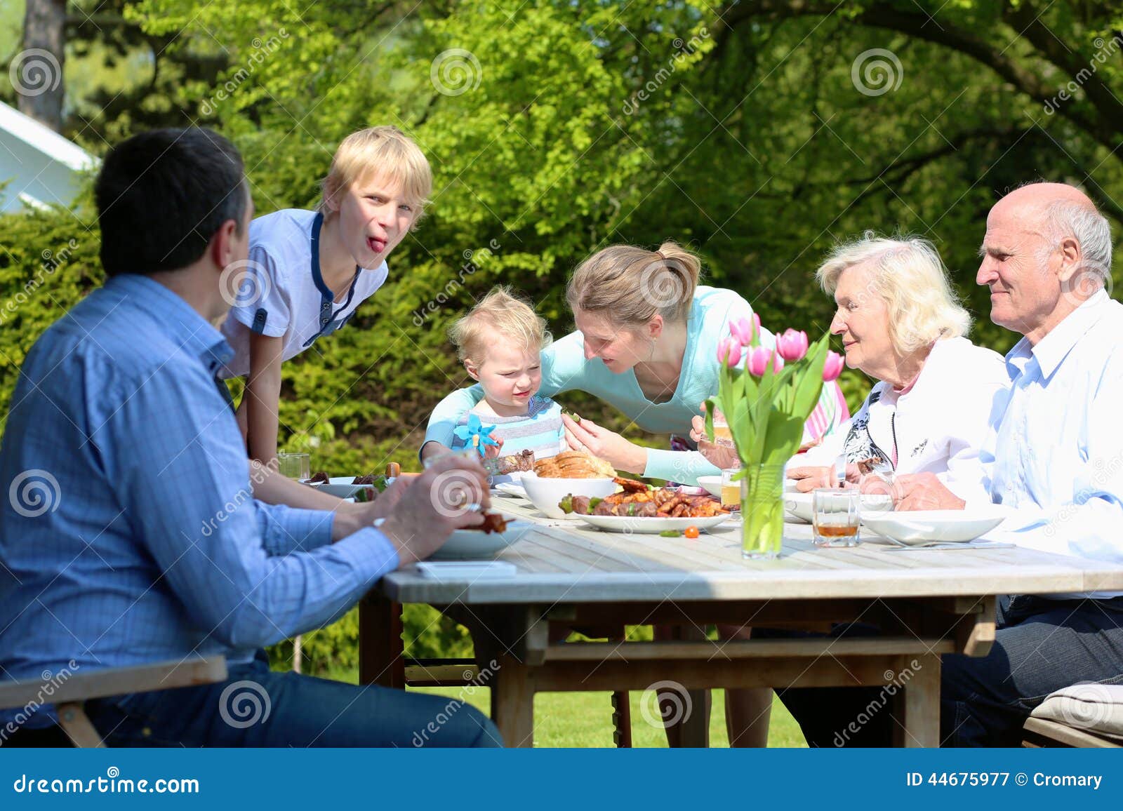 Grote familie die lunch in de tuin hebben. Grote gelukkige familie met jonge geitjes en grootouders die gezonde smakelijke bbq lunch hebben of diner in openlucht in de tuin bij de binnenplaats van hun huis die geroosterd vlees met groenten op een zonnige de zomerdag eten