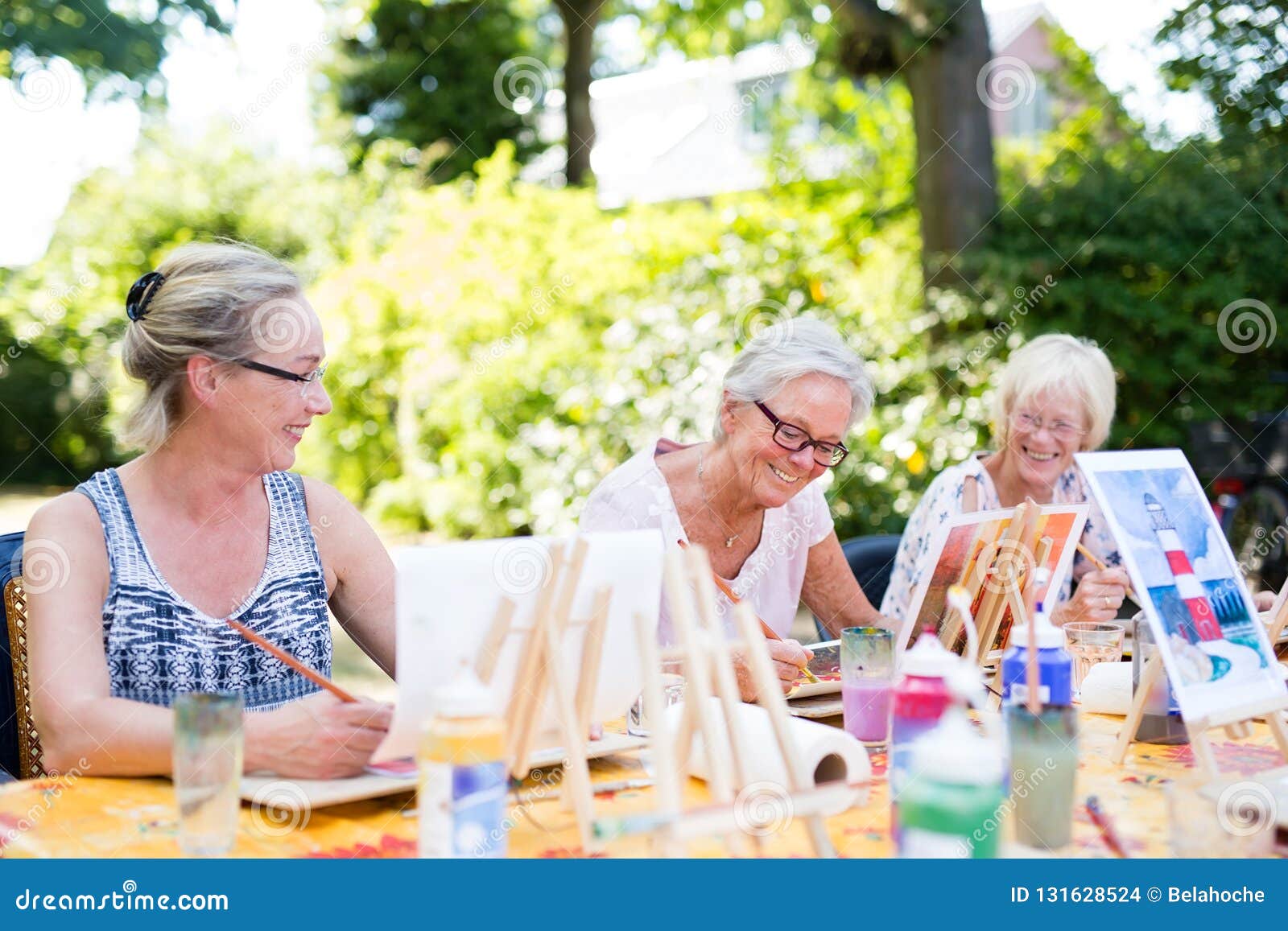 group of happy elderly women attending an outdoor art class in a garden or park painting from sample pictures on easels while