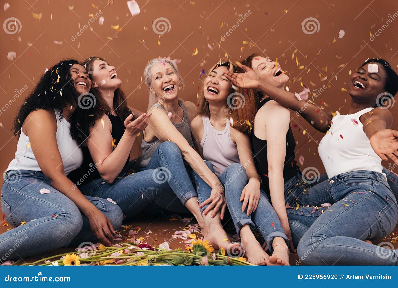 group of six laughing women of different ages sitting under falling flower petals. multi-ethnic smiling females having fun in