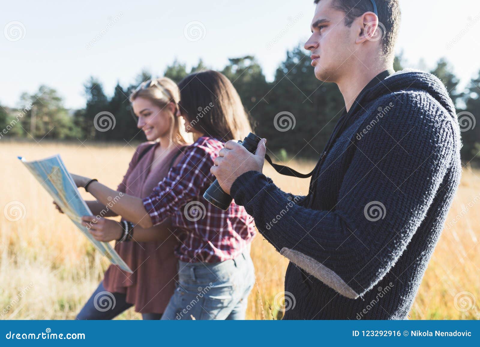 Gruppe junge Leute, die beim mounatin Wandern genießen. Junges Freundgenießen im Freien Zwei Mädchen, die im tiefen Gras stehen, Karte lächeln und betrachten, um besten Weg zu finden während junge Männer, die durch Ferngläser schauen