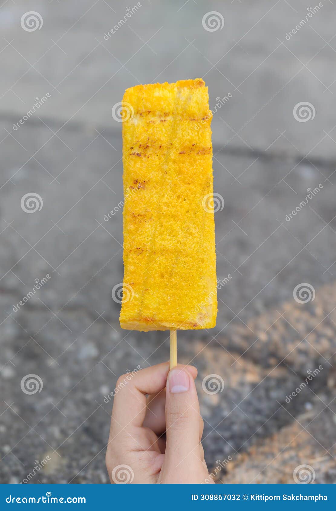 hand holding bread or yellow toast with slight burn marks on a wooden skewer on a vertical blurred gray