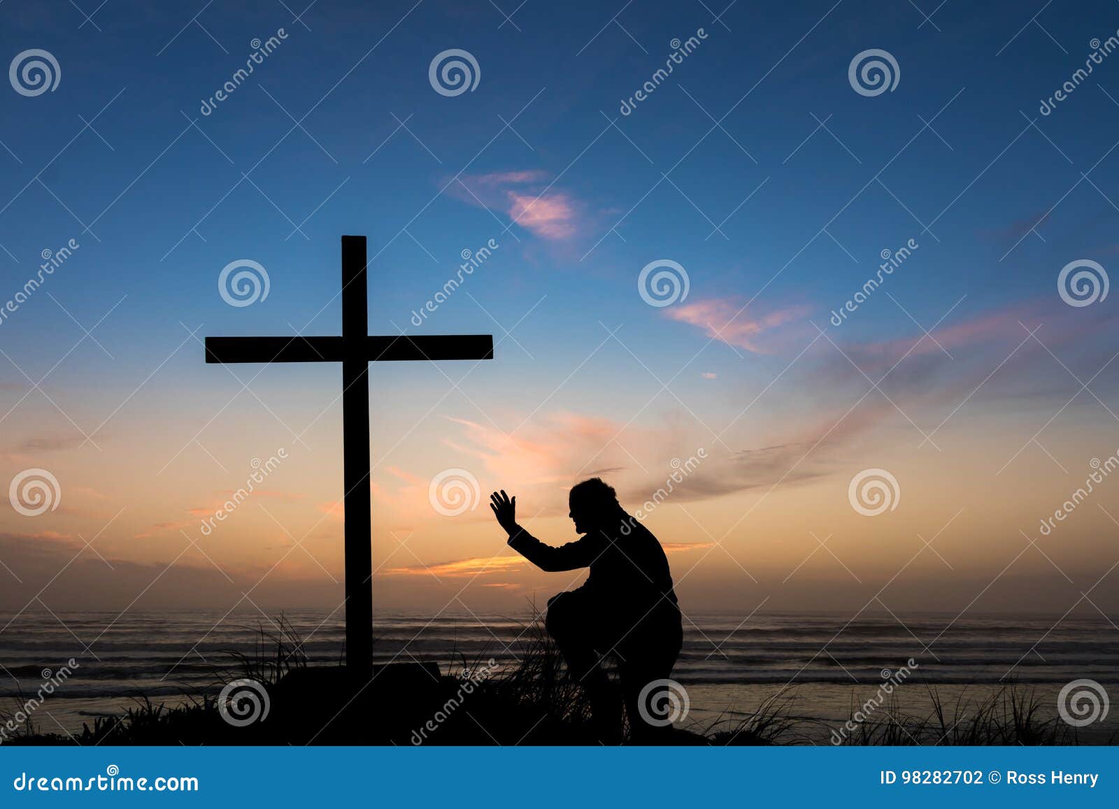 Hand to Salvation. Man praying with his hand up by a black cross on a beach with a dusk sky sunset behind it.