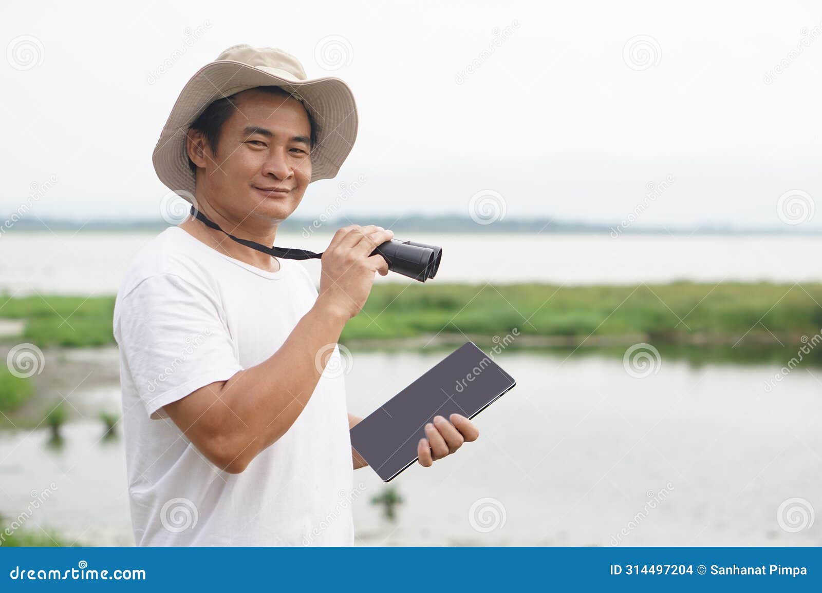 handsome asian man ecologist is surveying nature at the lake, holds binoculars and smart tablet.