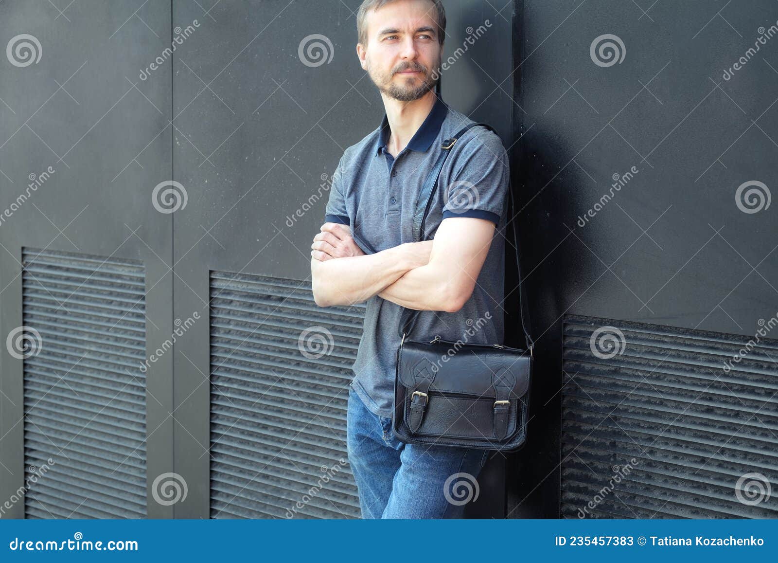 handsome bearded millennial casual wearing man with stylish leather bag standing near black wall in the city streeet