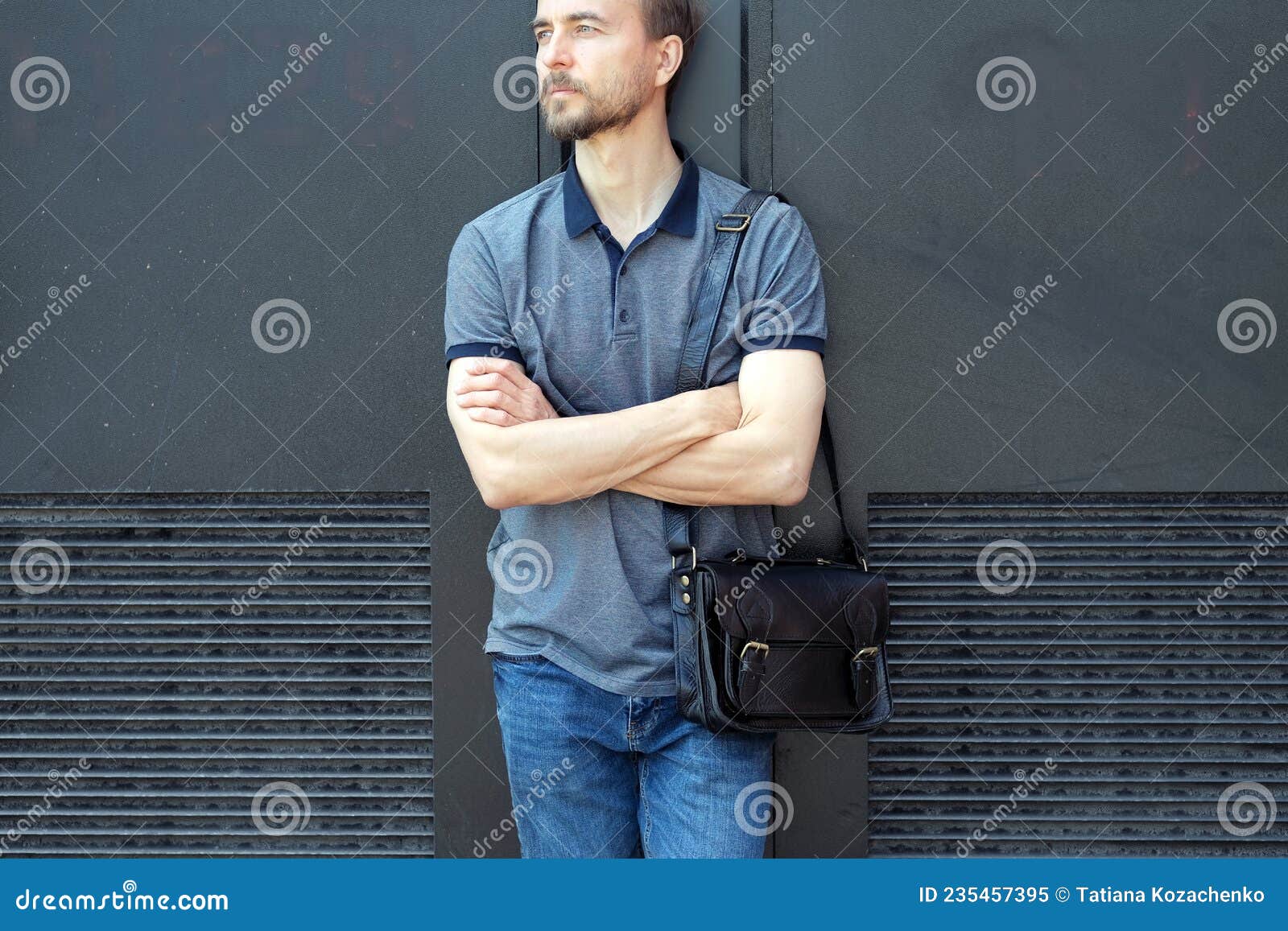 handsome bearded millennial man in polo shirt and jeans with stylish leather bag standing near black wall in the city streeet