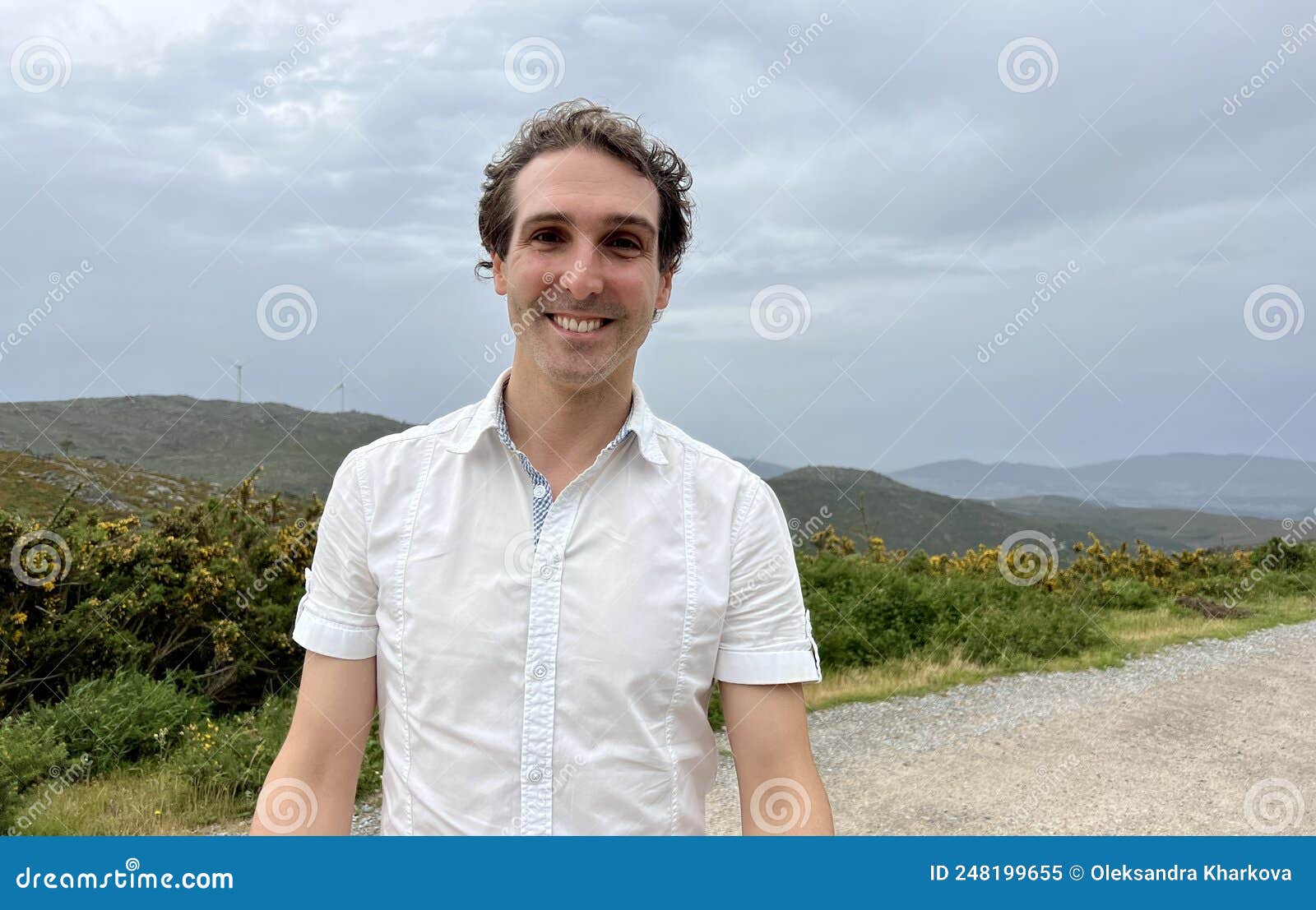 a handsome french or spaniard man in a white shirt stands against the backdrop of mountains