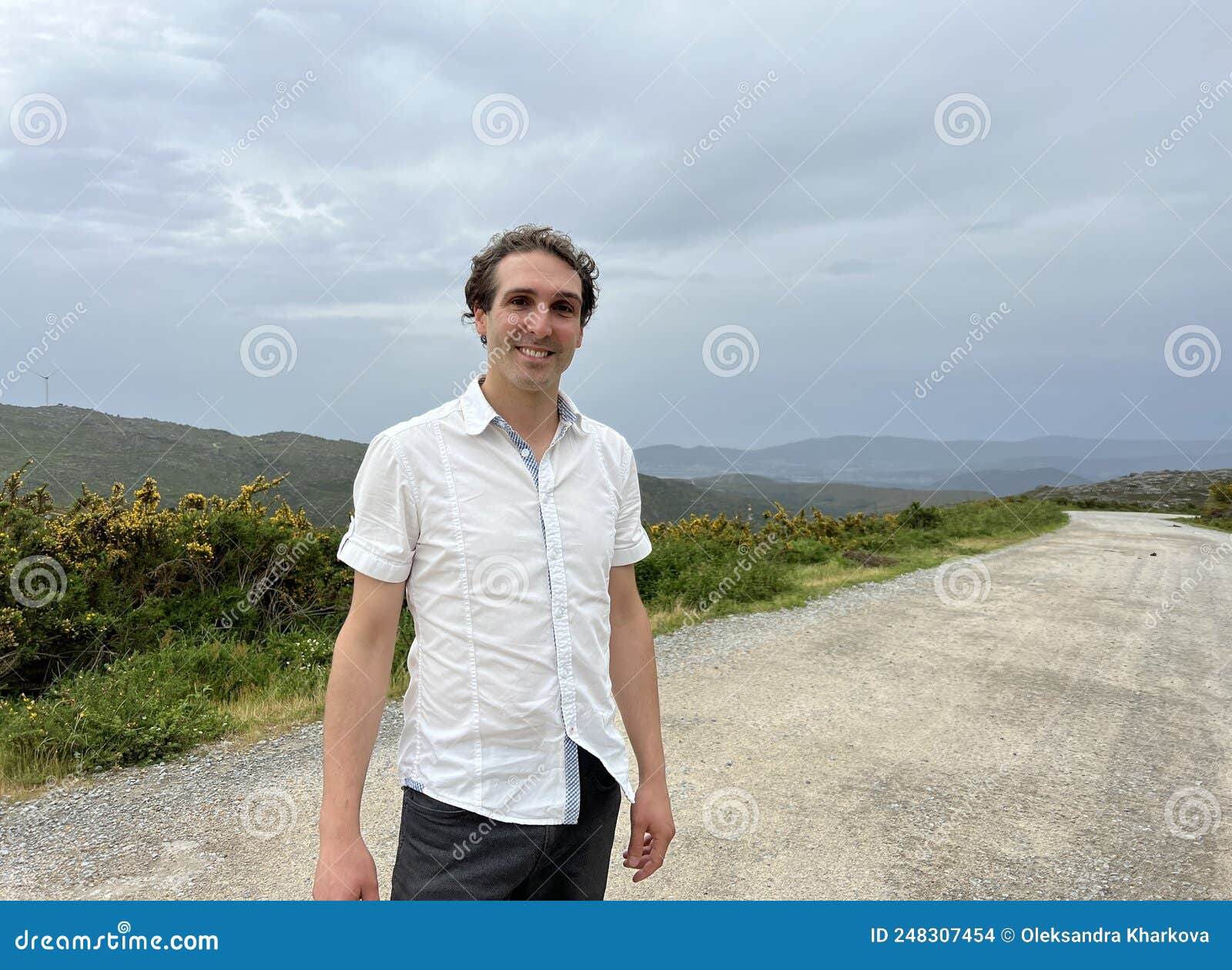a handsome french or spaniard man in a white shirt stands against the backdrop of mountains