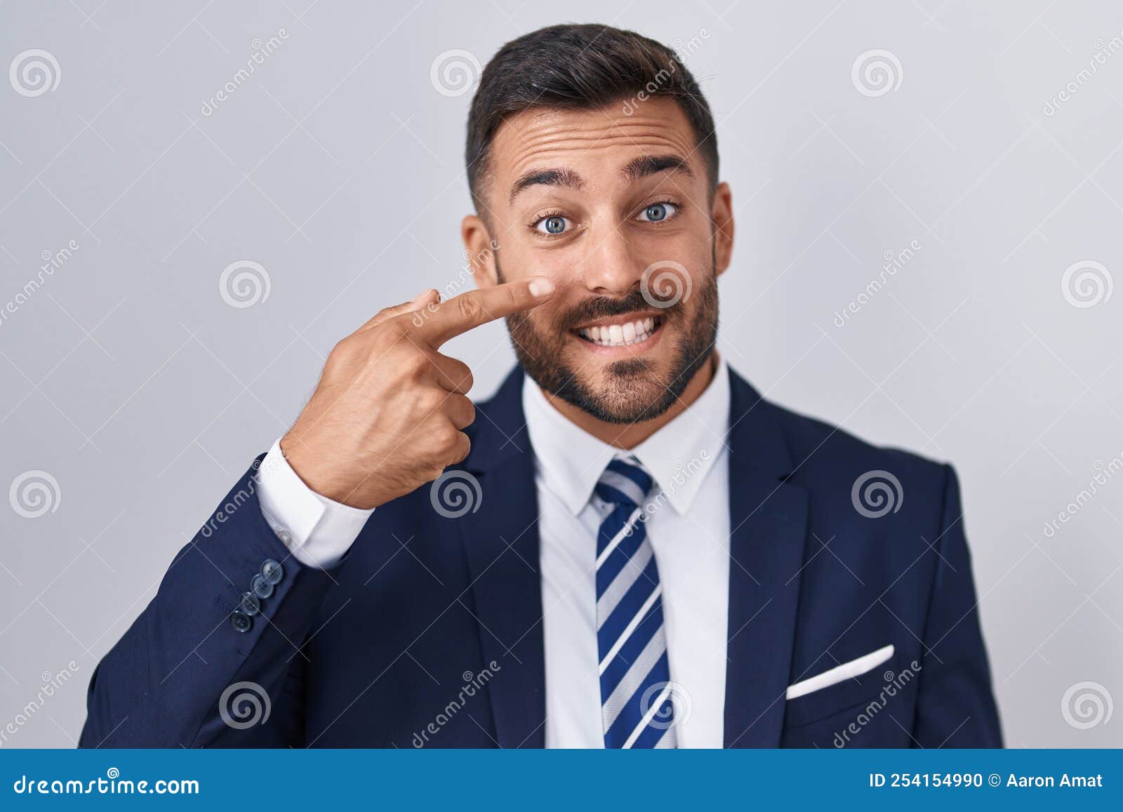 Handsome Hispanic Man Wearing Suit And Tie Pointing With Hand Finger To Face And Nose Smiling