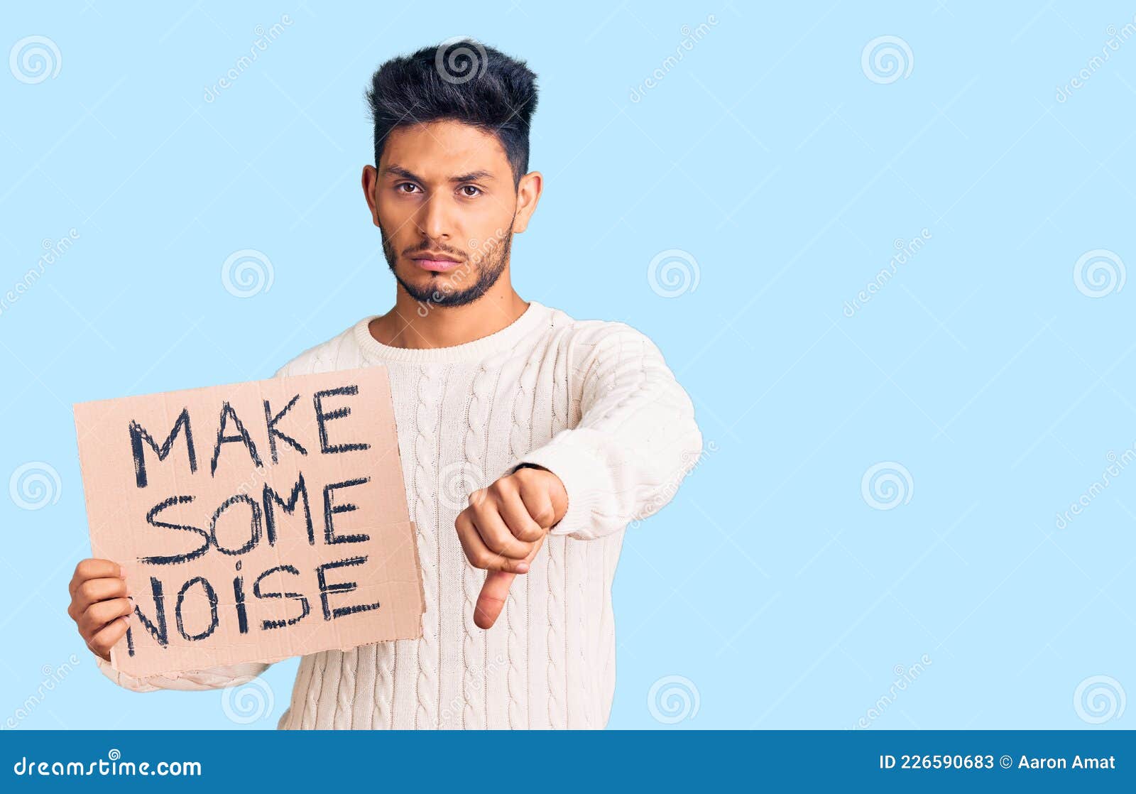 handsome latin american young man holding make some noise banner with angry face, negative sign showing dislike with thumbs down,