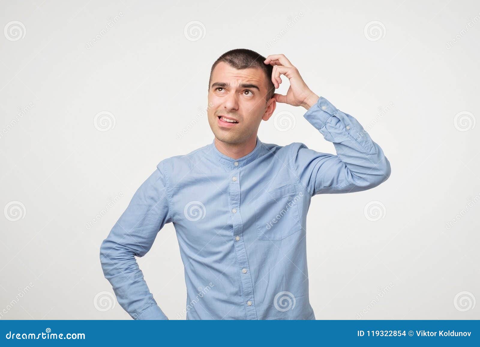 handsome spanish guy in blue shirt, frowning and looking unsatisfied while scratching head