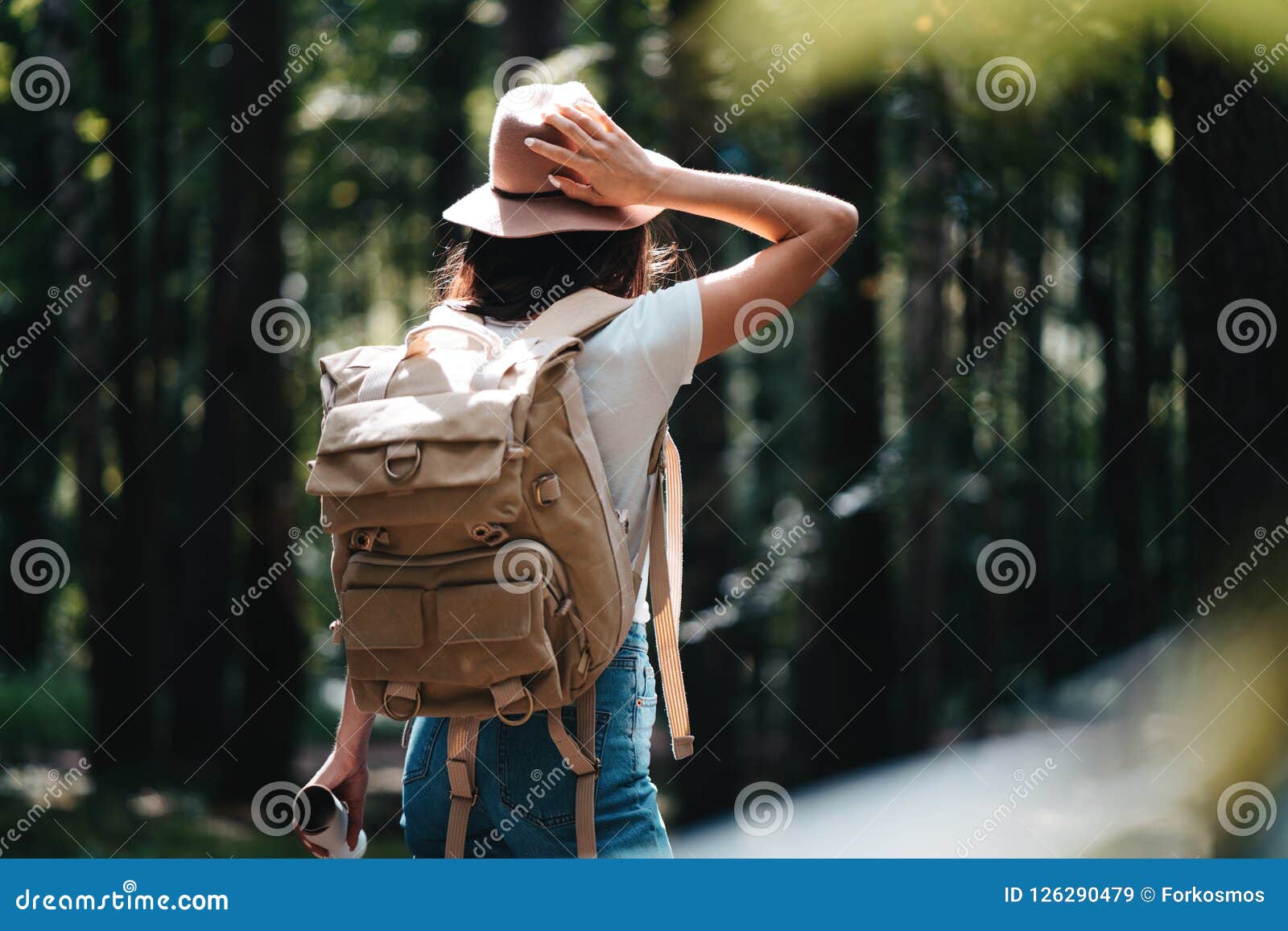 handsome traveler woman with backpack and hat standing in forest. young hipster girl walking among trees on sunset