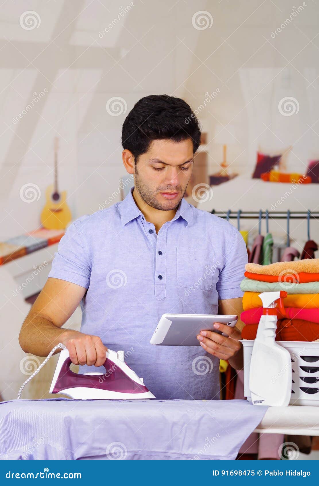 Handsome Young Man Ironing Clothes on Ironing Board while he is Using