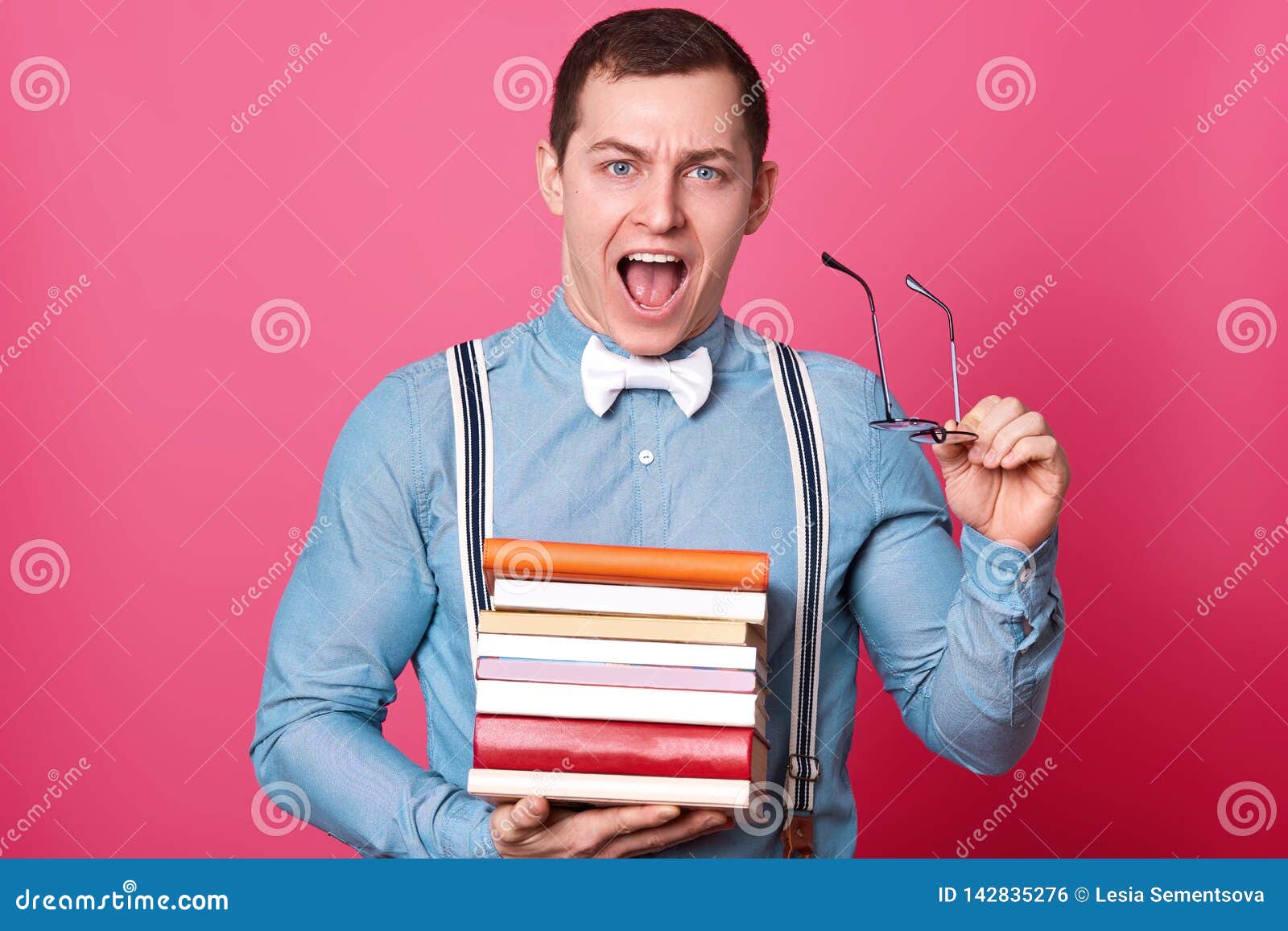 handsome young man with stack of books looking at camera and screaming while standing on rosy background. tired student finds out