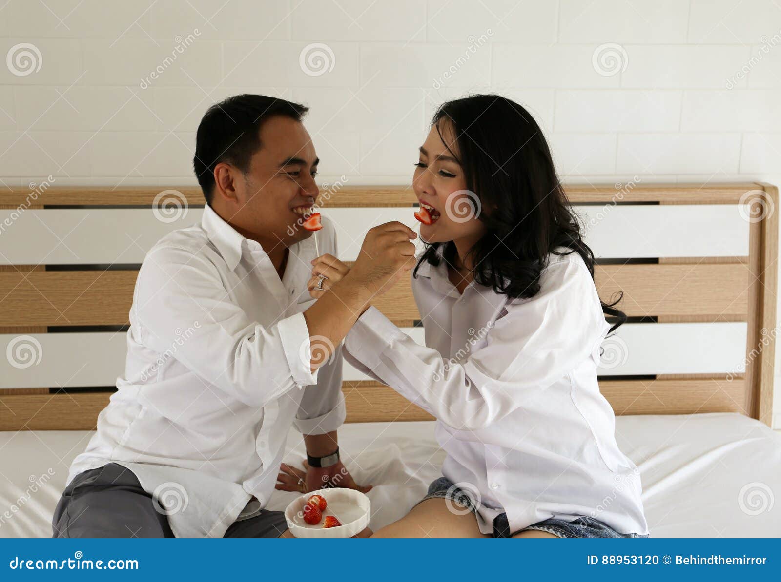Happy Asian Couple In White Shirt Feed Each Other Fruit On The Bed