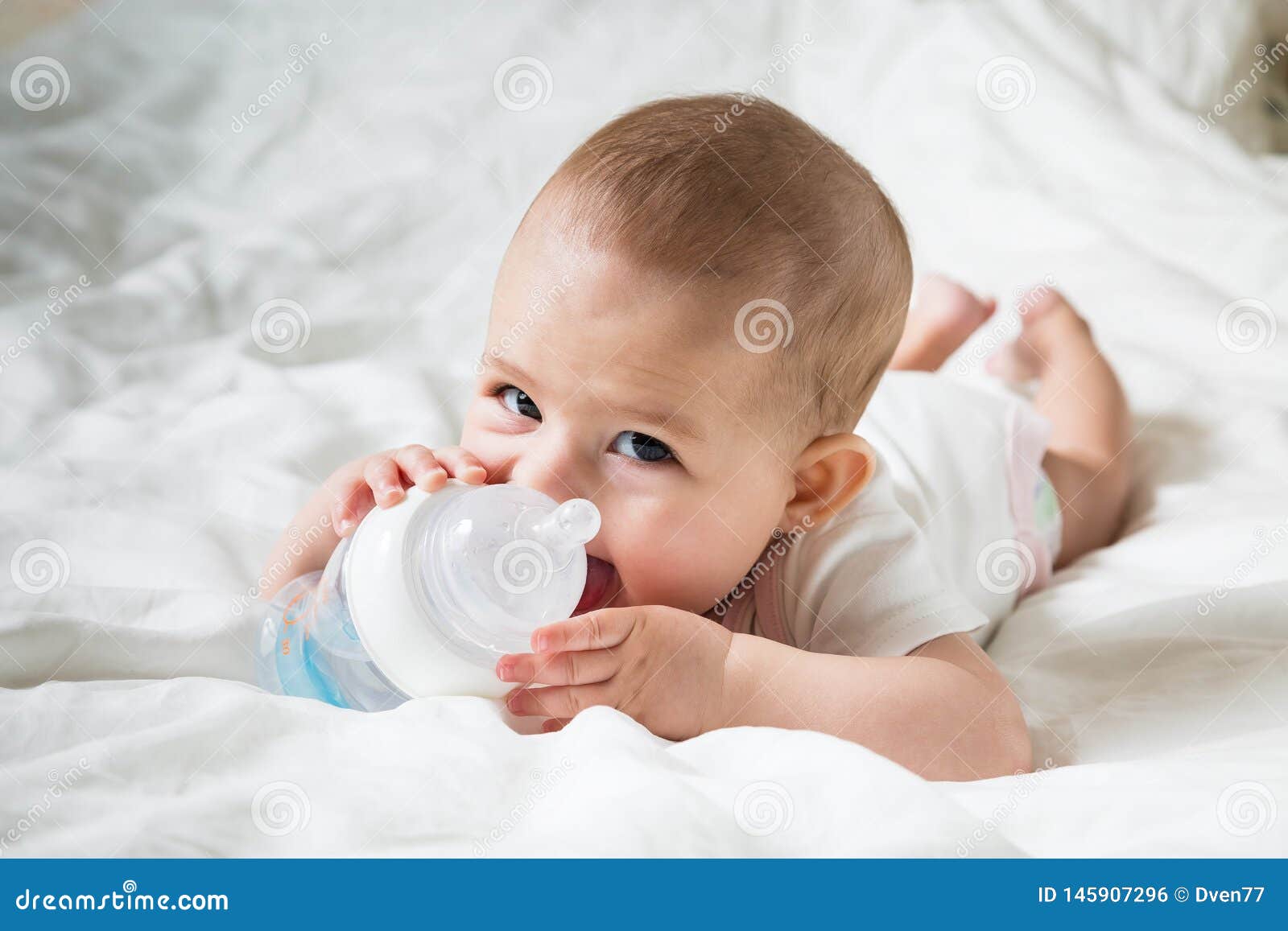 happy baby in white clothes on a white bed. drink water from the special bottle. close up, infant leer. tricky glance
