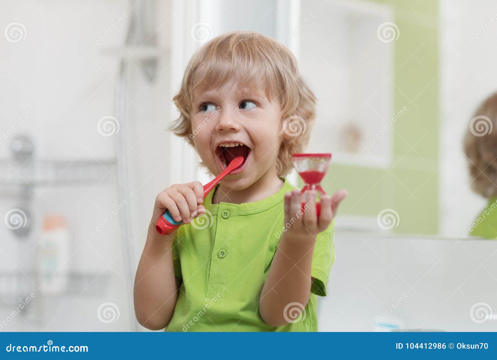 happy child brushing teeth near mirror in bathroom. he is monitoring lasting of cleaning action with hourglass.