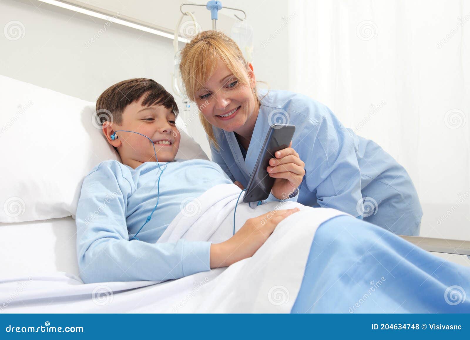 happy child lying in bed in hospital room and smiling nurse using smartphone surfs the internet wearing earphones