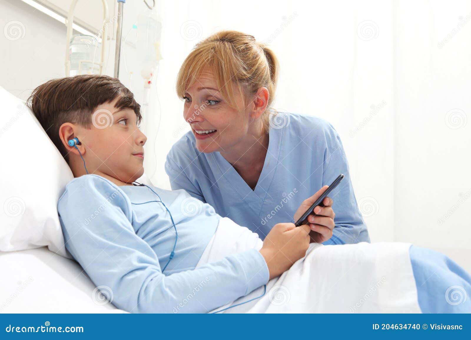 happy child lying in bed in hospital room and smiling nurse using smartphone surfs the internet wearing earphones