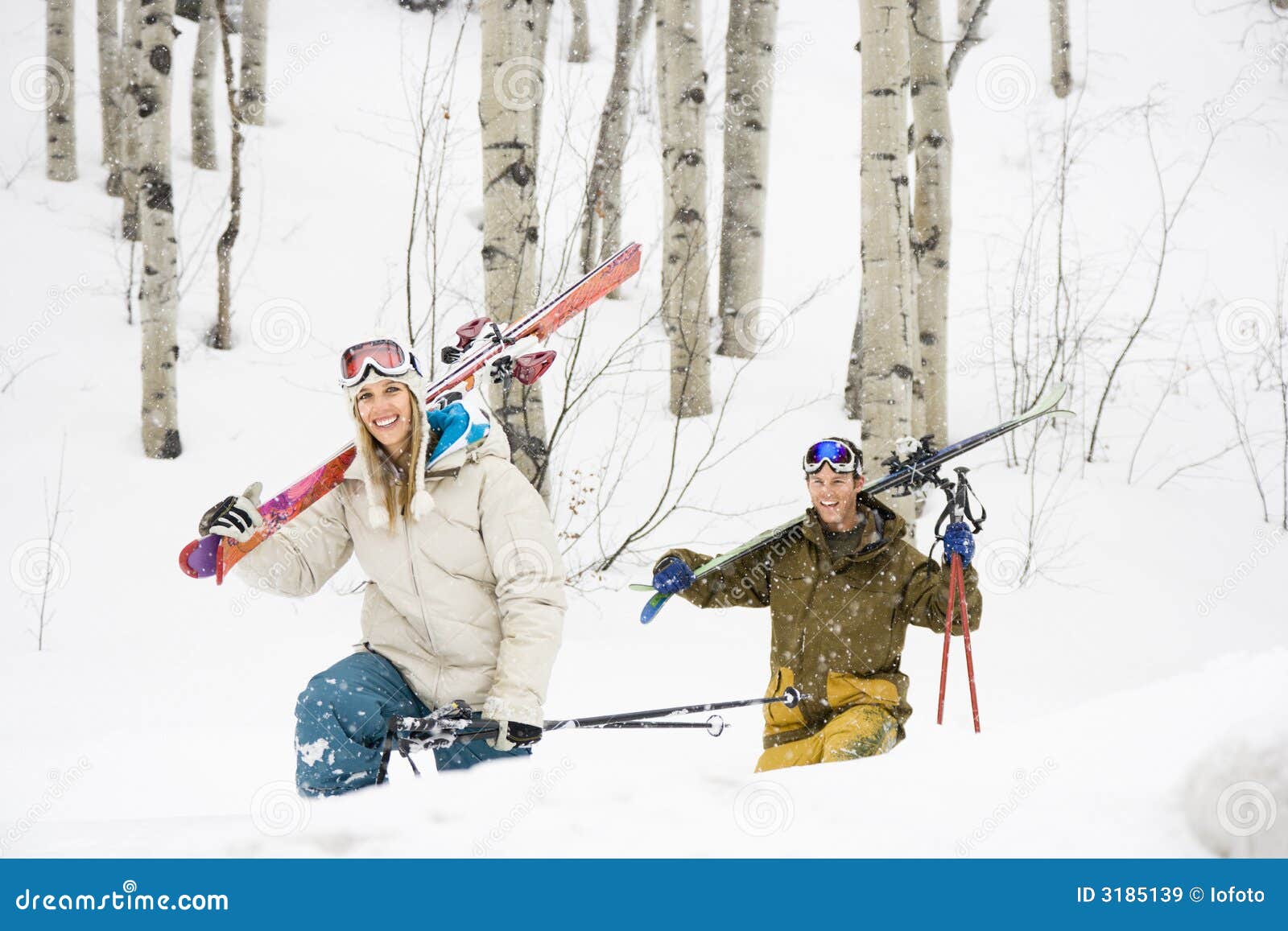 Happy couple on ski vacation. Happy young couple walking through snow carrying skis over shoulders smiling.