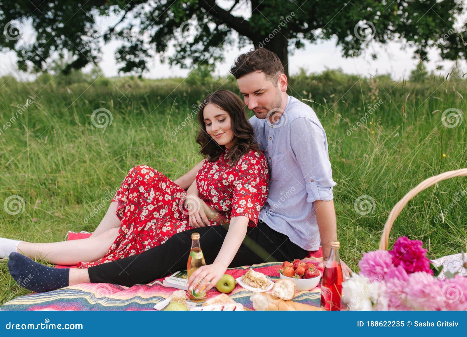 happy couple spend time outdoors on mini picnic. attractive woman and handsome man sits on red blanker by the big tree