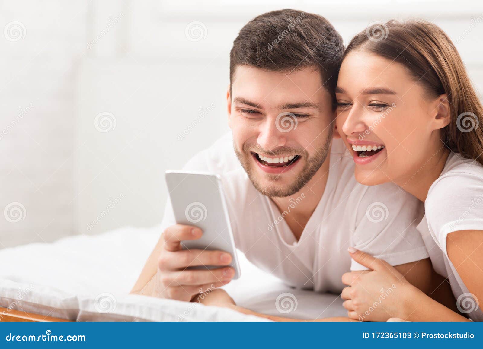 Happy Couple Using Smartphone Texting Lying in Bed in Bedroom Stock