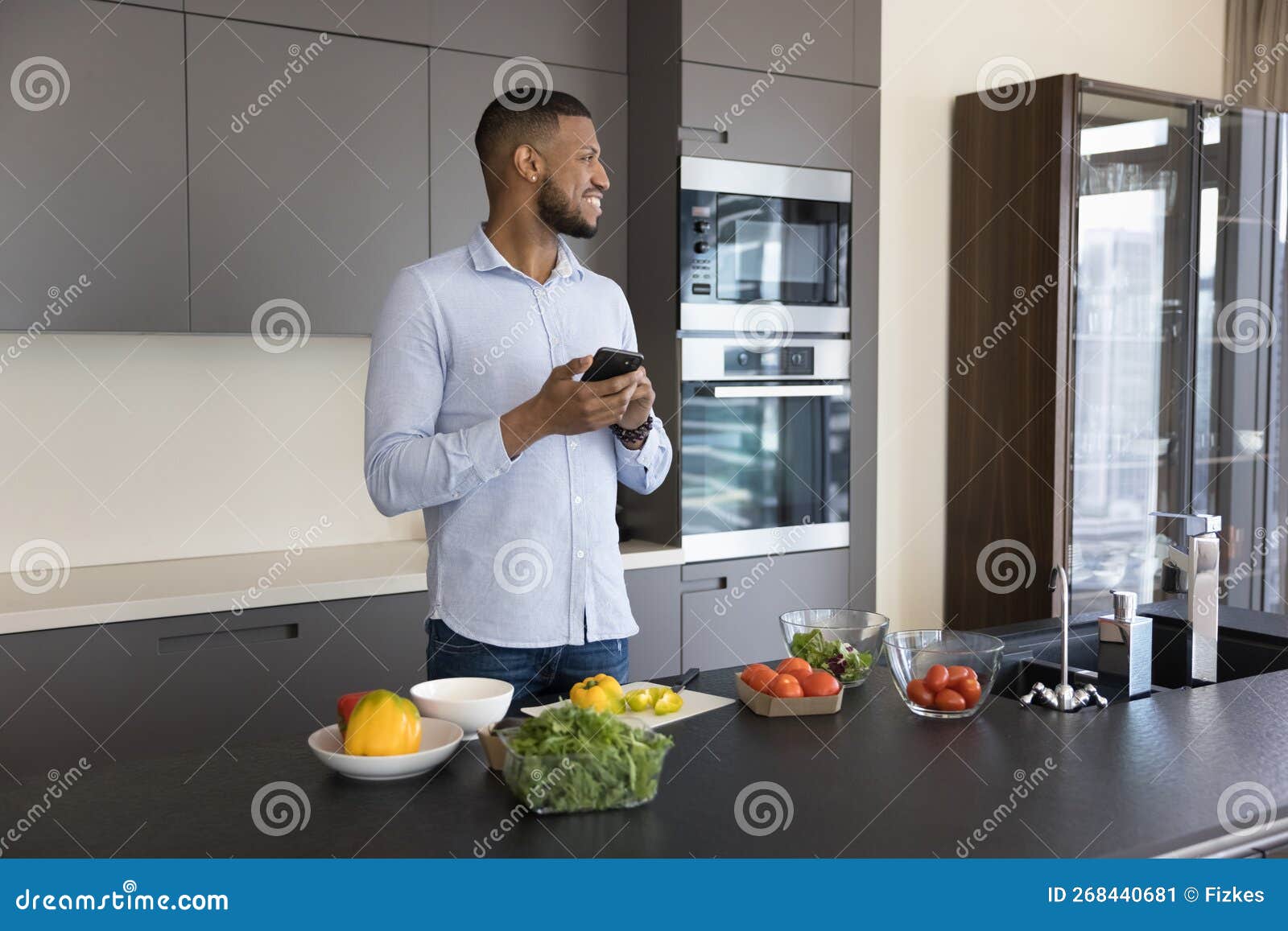 Happy Dreamy Young Black Guy Using Mobile Phone in Kitchen Stock Image