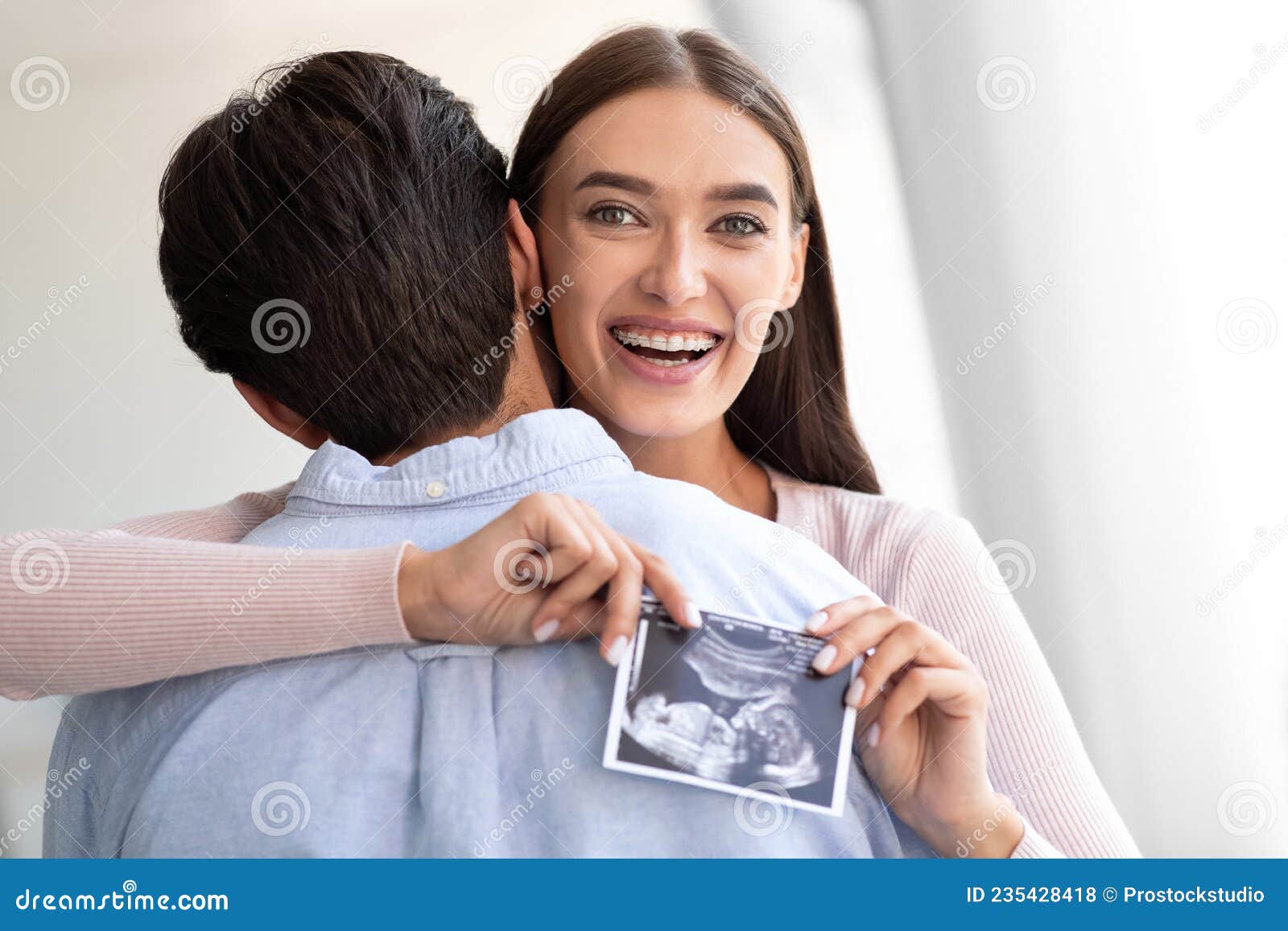 Happy European Young Wife Hugs Husband and Holds Ultrasound Picture of Baby in Living Room Interior Stock Photo