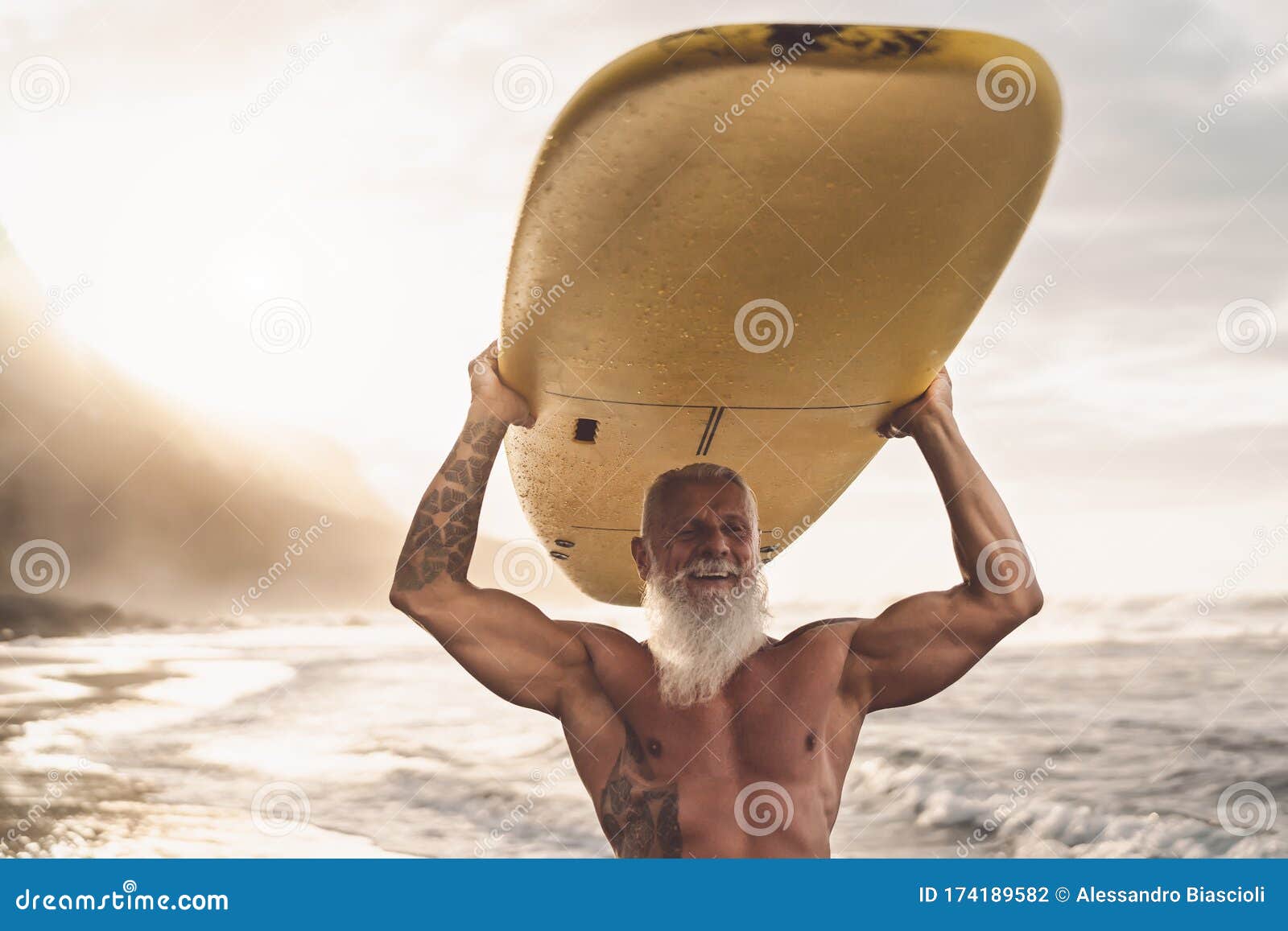 happy fit senior having fun surfing at sunset time - sporty bearded man training with surfboard on the beach