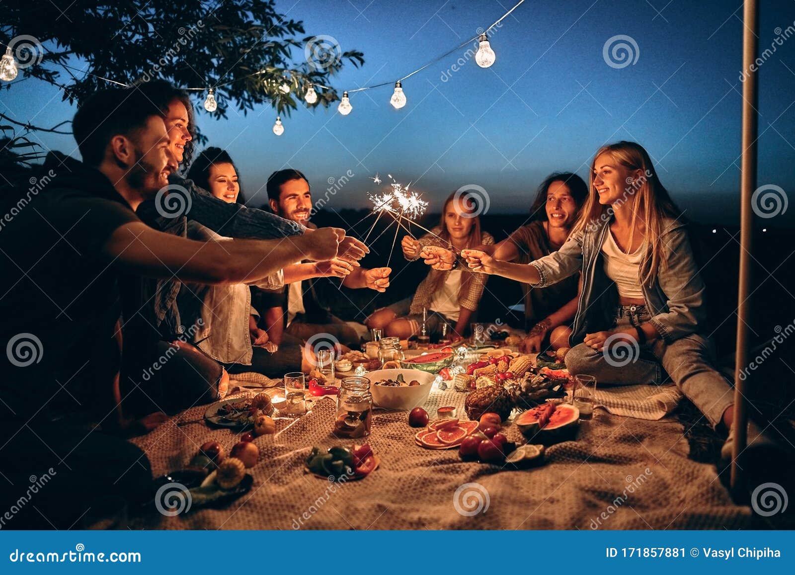 happy friends having fun with fire sparkles. young people millennials camping at picnic after sunset