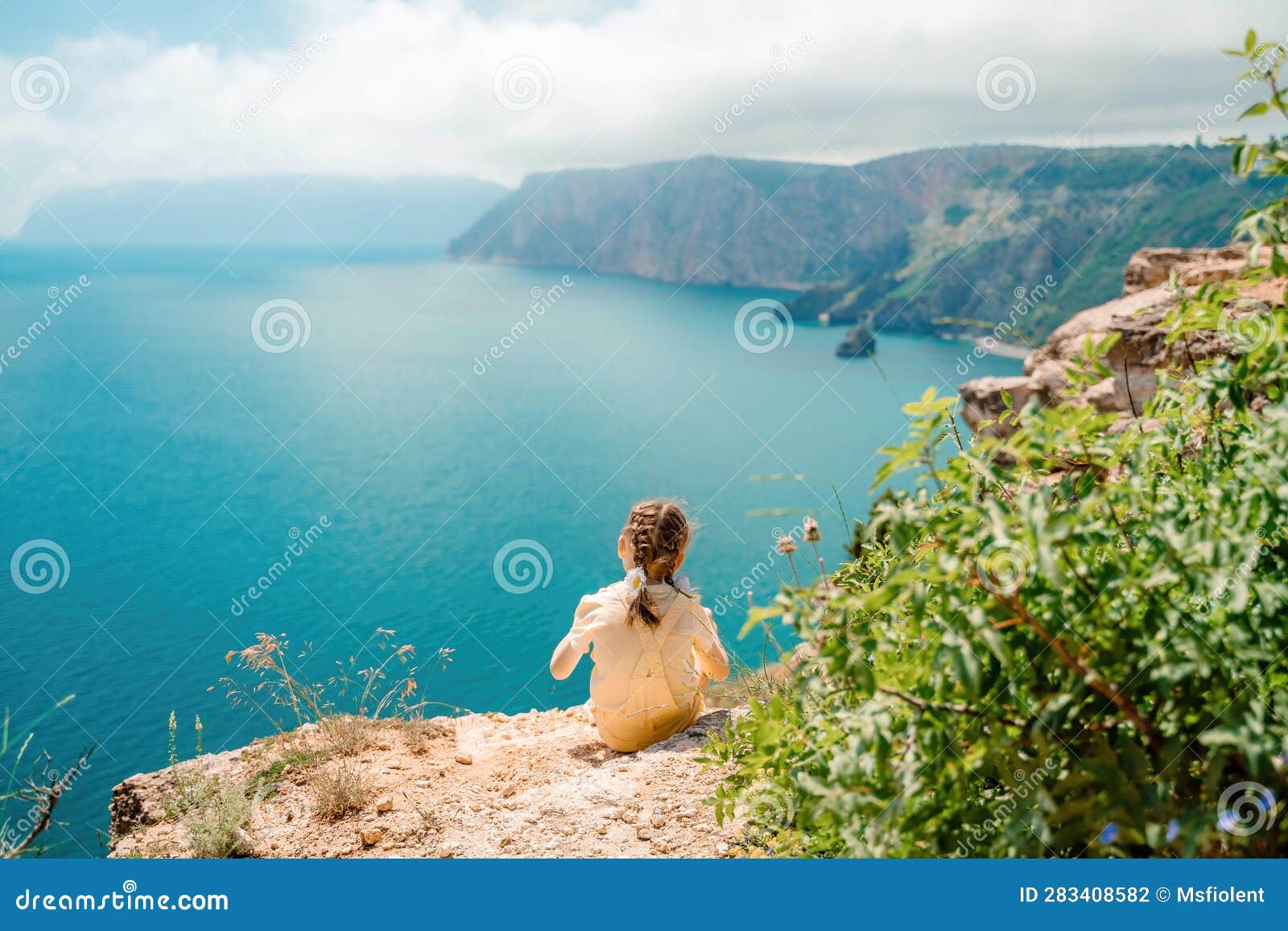 Happy Girl Perched Atop A High Rock Above The Sea Wearing A Yellow Jumpsuit And Braided Hair