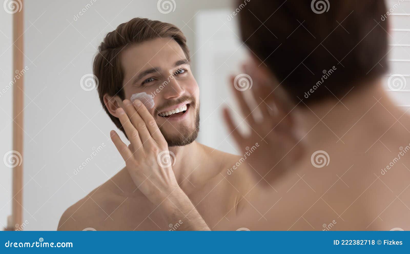 happy handsome metrosexual guy applying sunscreen on face at mirror