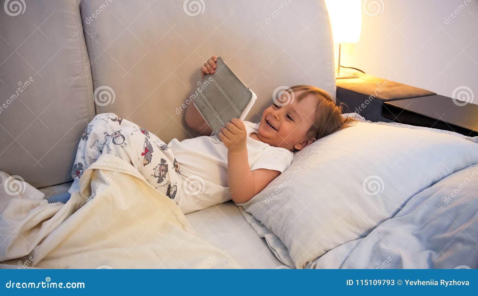 portrait of happy laughing toddler boy lying in bed with digital tablet
