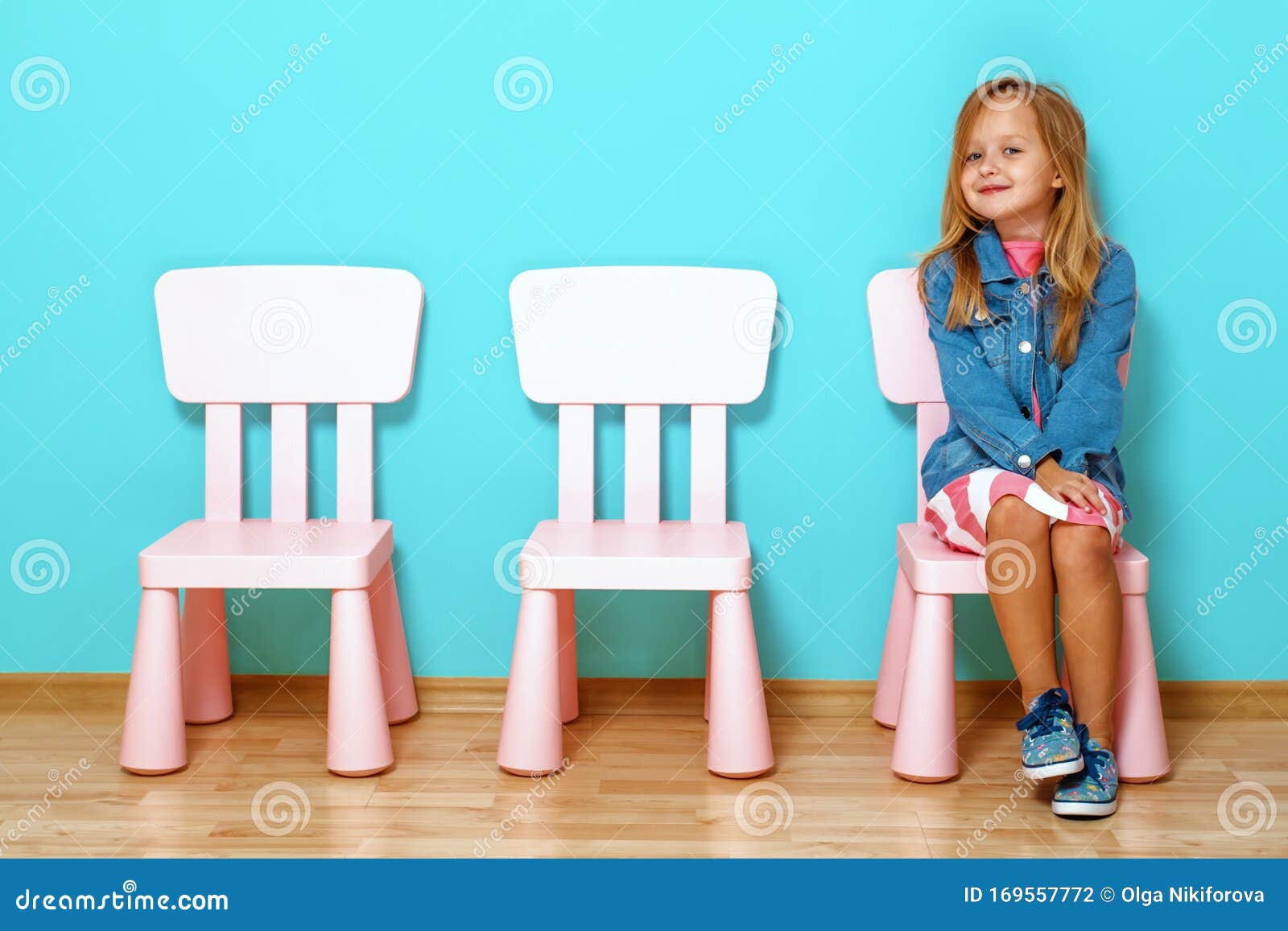 Happy Little Child Girl is Sitting on the Chair Against the Background of  the Blue Wall. Nearby are Empty Chairs Stock Photo - Image of funny,  background: 169557772