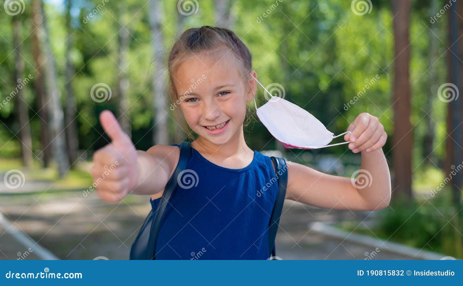 happy little girl takes off the mask and shows thumb outdoors. joyful smiling schoolgirl with a backpack pushes the mask