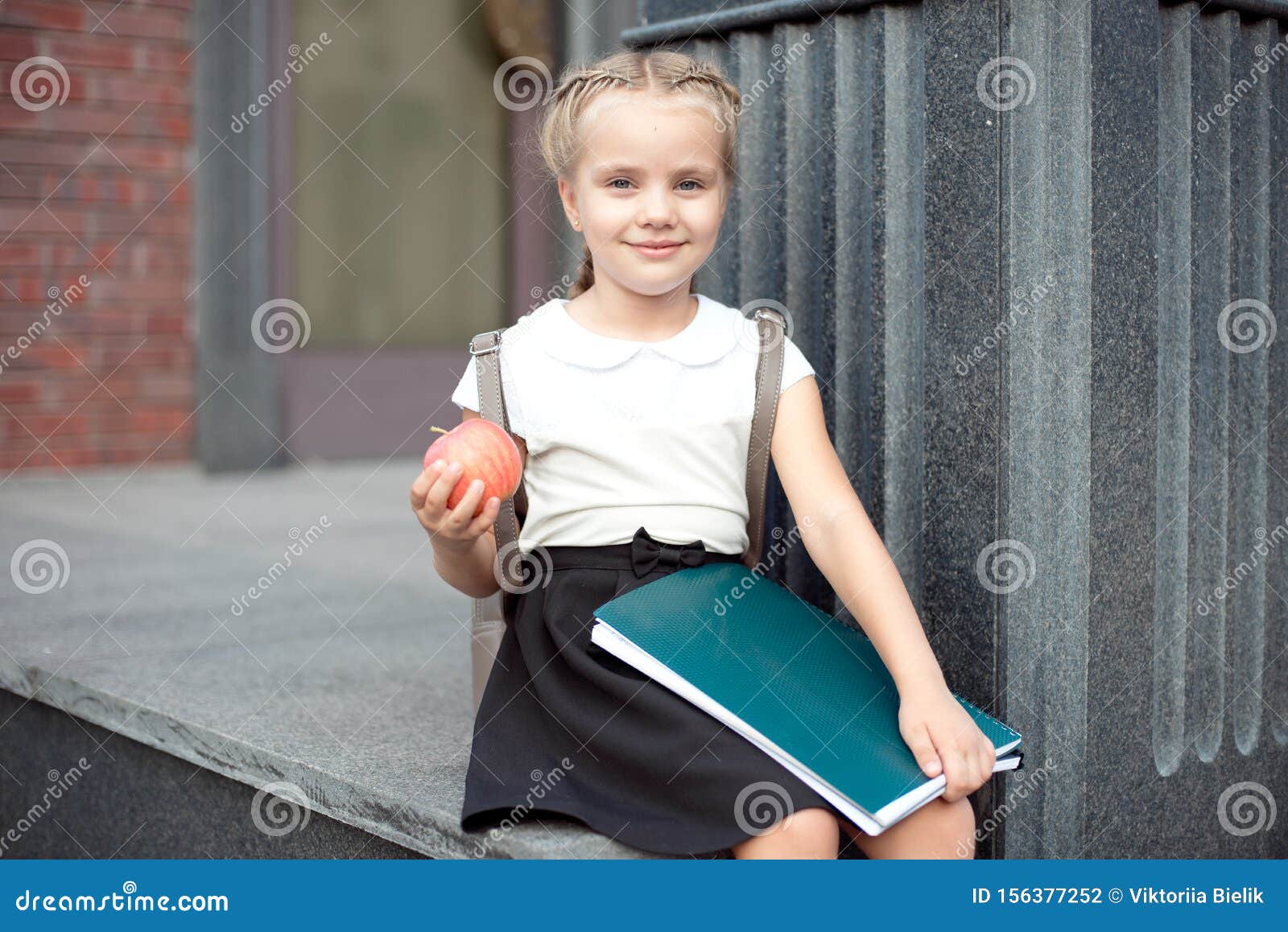 happy little schoolgirl with blond hair in school uniform with lunch, book sits near school