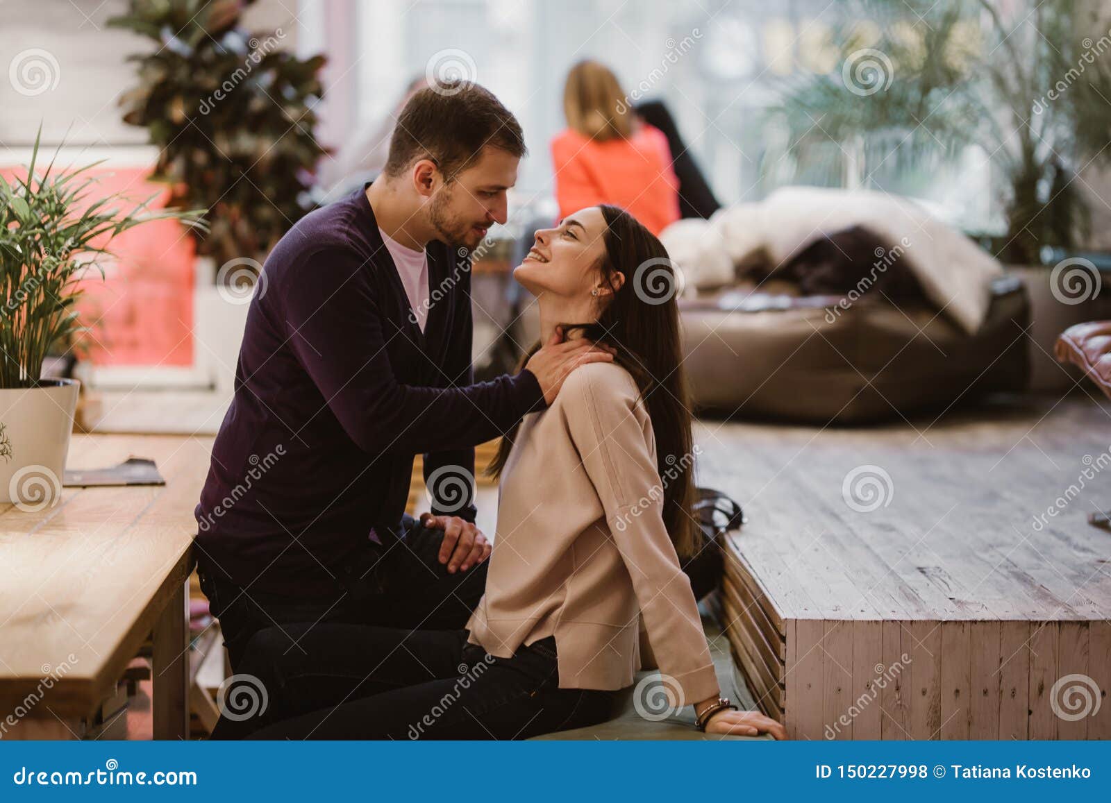 Happy Loving Guy Puts His Hands At The Girl`s Shoulders Sitting At The Table In The Cafe And