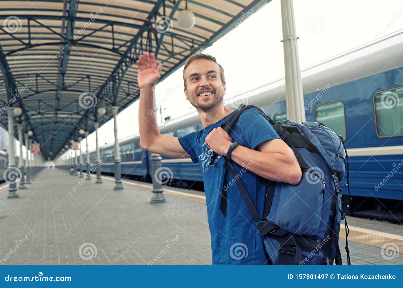 happy man tourist with backpack stand on railway station platform, greeting friends or saying goodbye, waving his hand. travel by