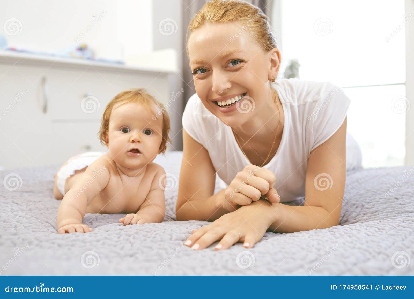 Happy Mother Smiles at Baby Sitting on Bed in Room. Stock Image - Image ...