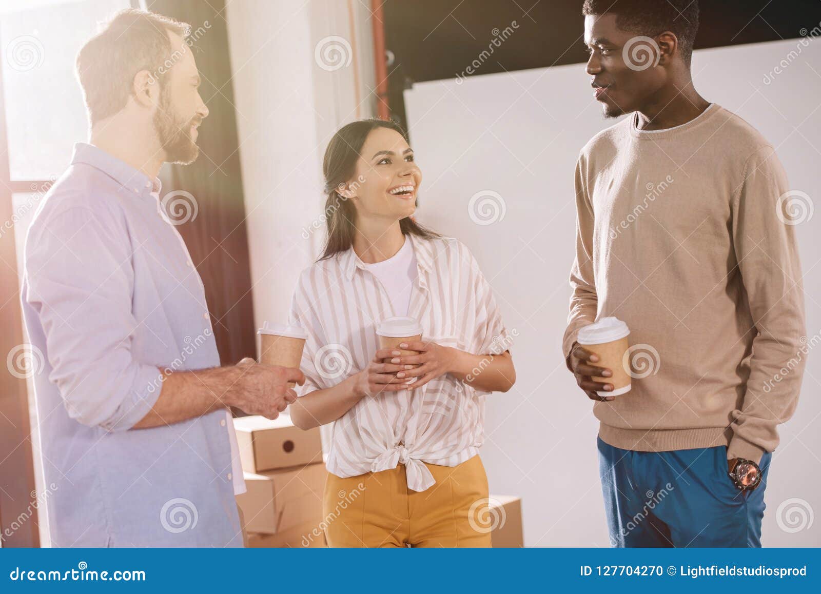 happy multiethnic coworkers holding paper cups and talking in new office