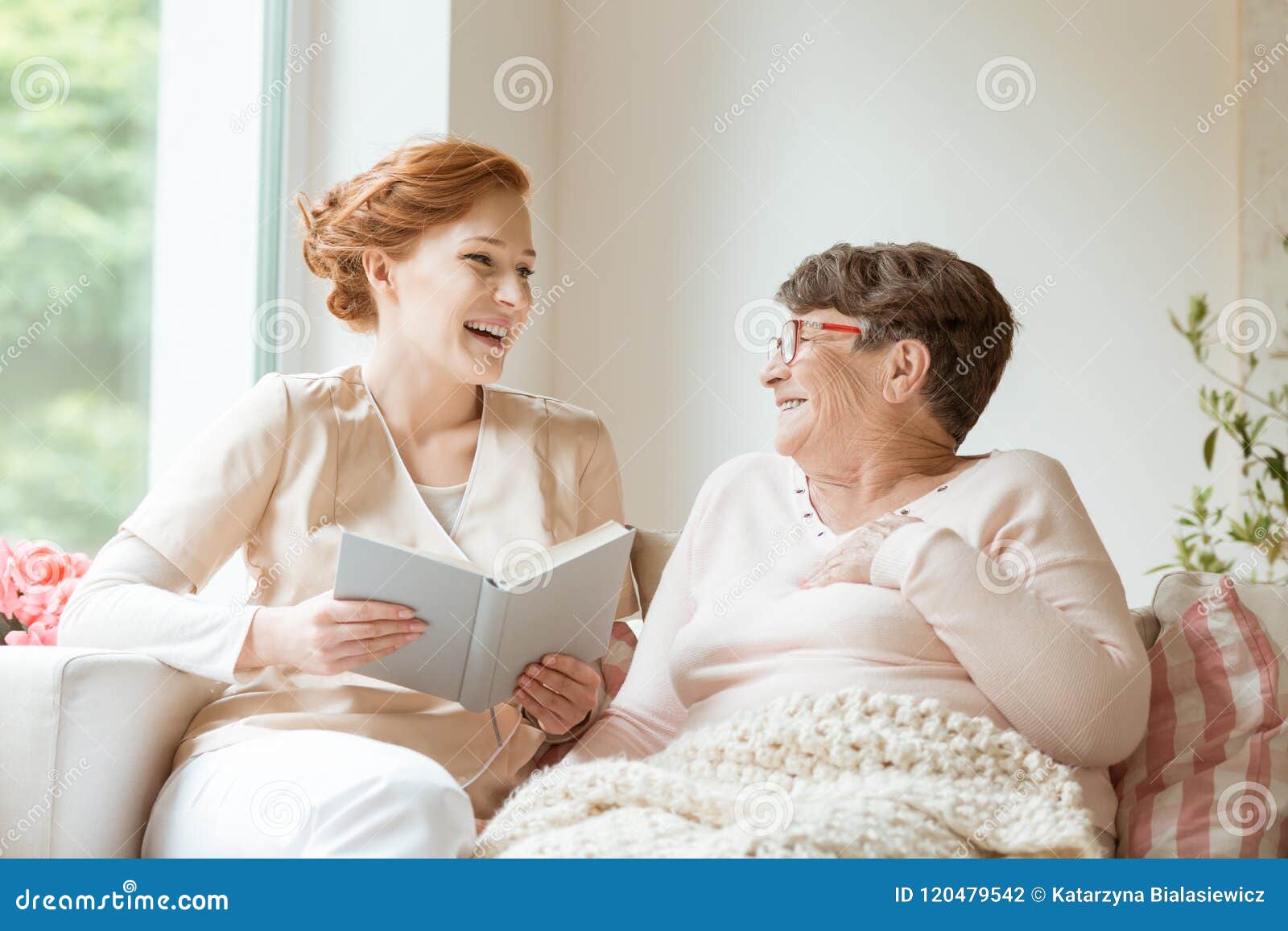happy nurse reading a funny book to her elderly patient in a private nursing home