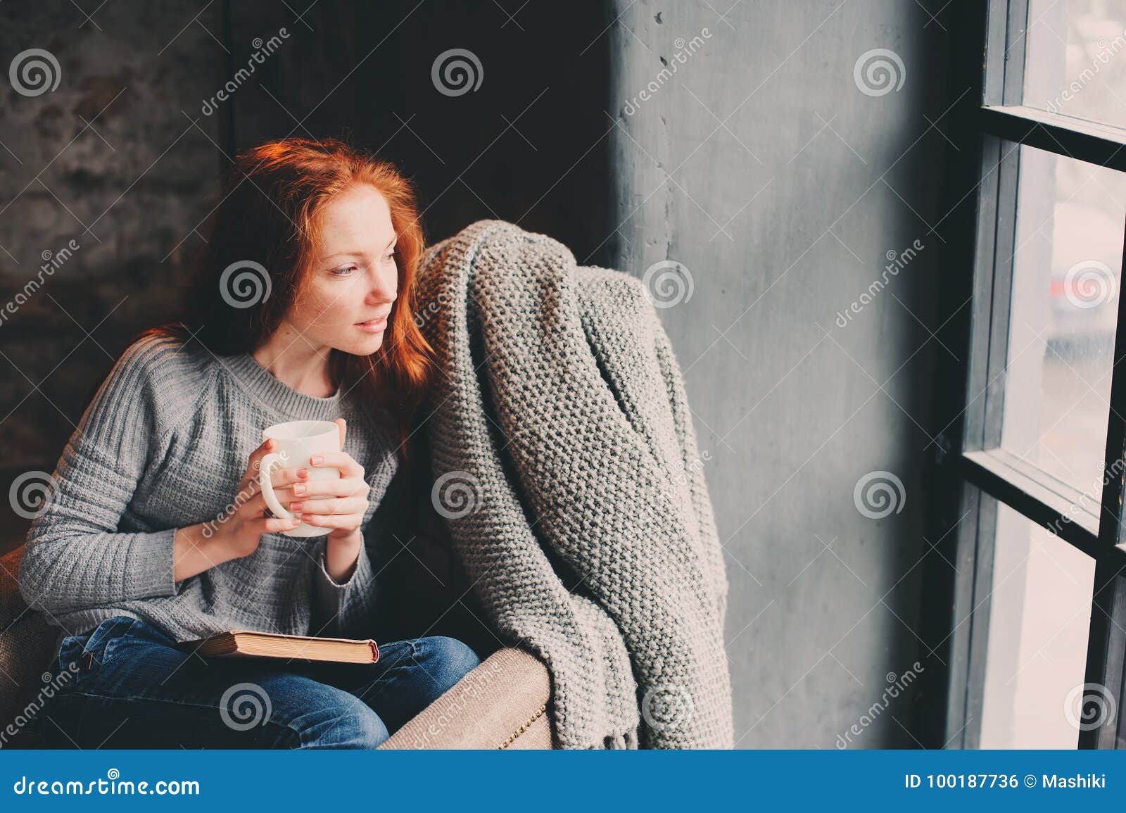 happy redhead woman relaxing at home in cozy winter or autumn weekend with book and cup of hot tea, sitting in soft chair