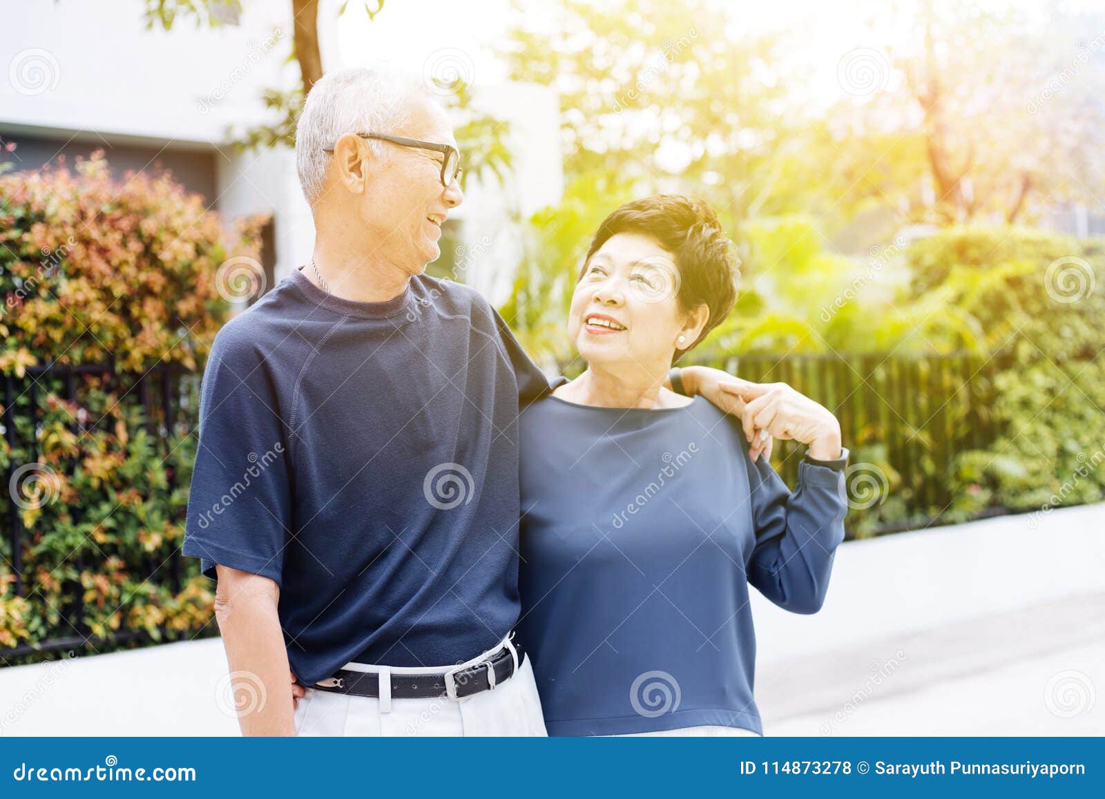 happy retired senior asian couple walking and looking at each other with romance in outdoor park and house in background.