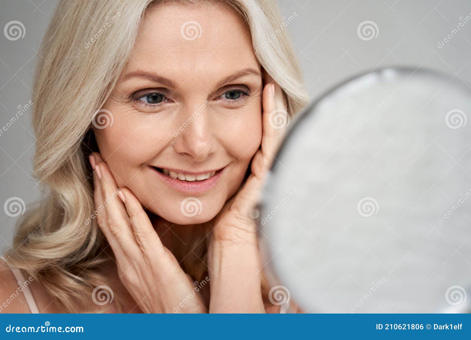 happy 50s middle aged woman touching face skin looking in mirror.